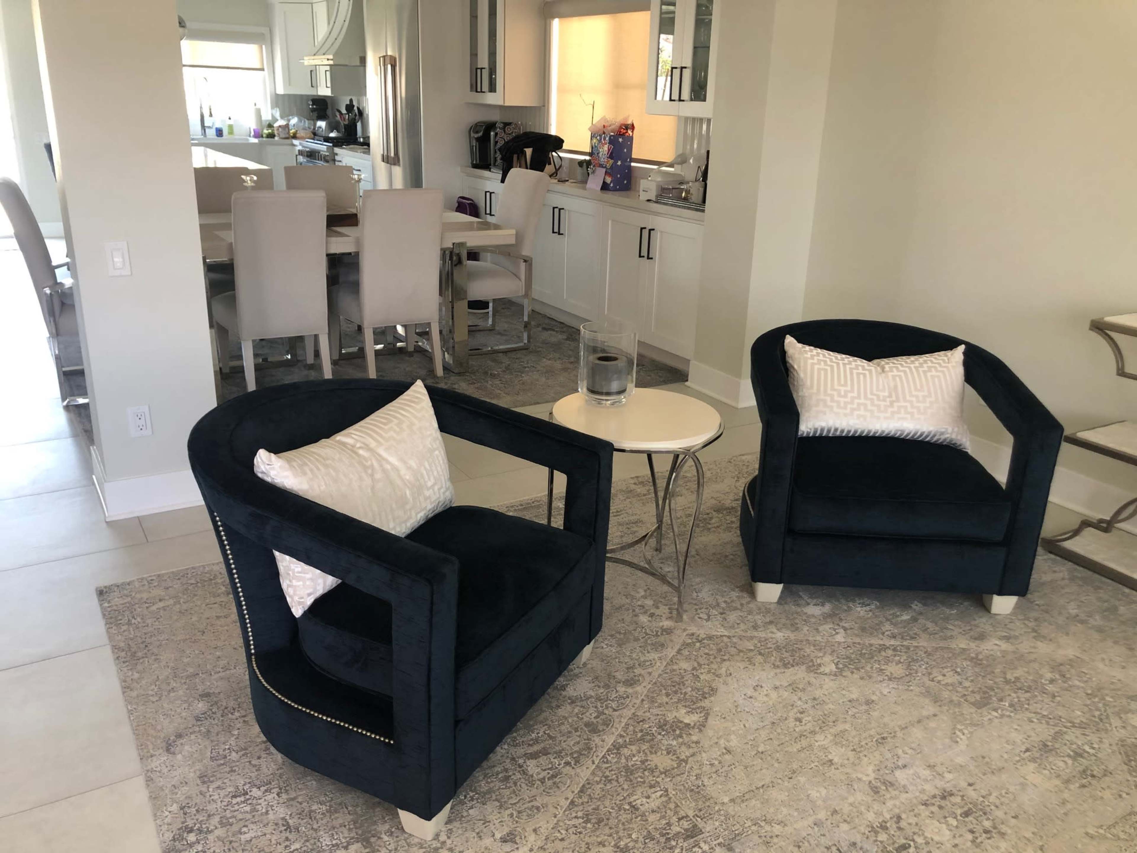 The image shows two dark blue upholstered chairs with decorative pillows positioned near a small round table in a modern, light-colored living space that leads to a dining area.