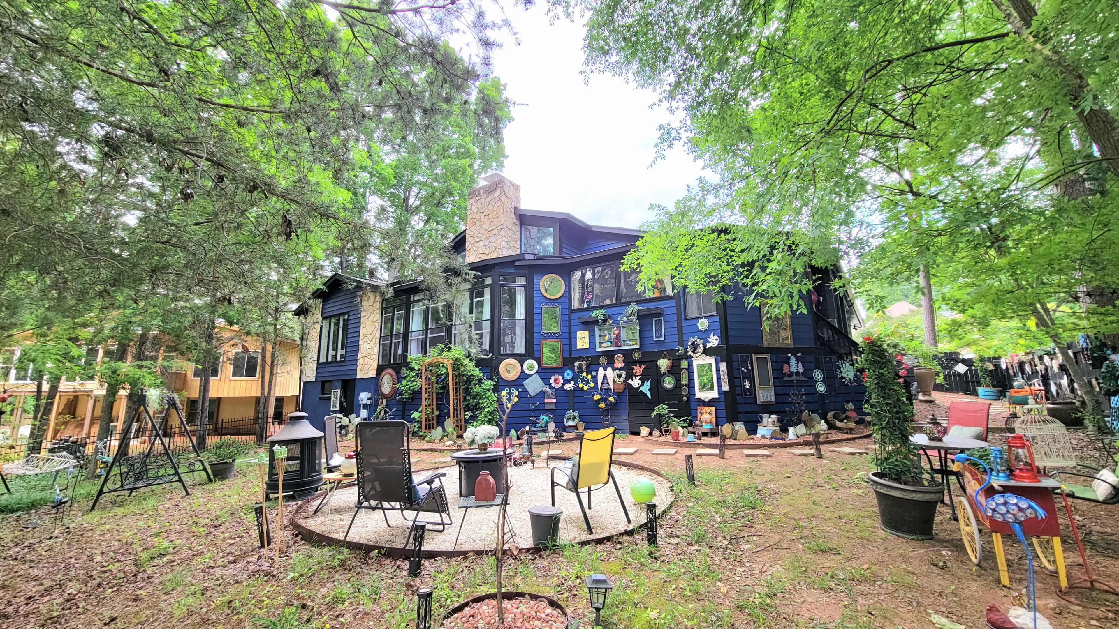 A large dark blue house surrounded by trees, with various decorations on its walls and a circular garden area featuring chairs and colorful items.