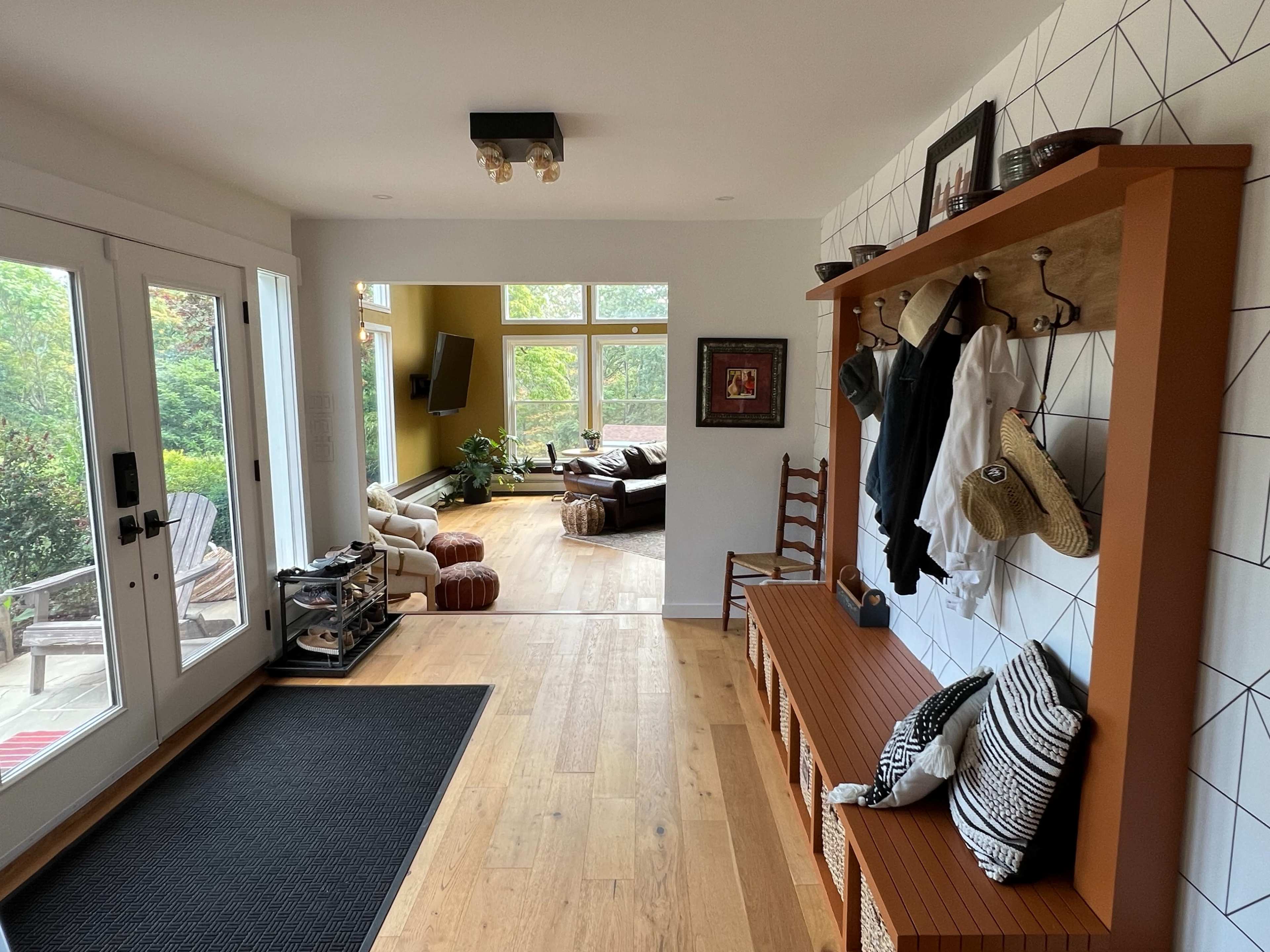 A hallway with a wooden bench, coat hooks, and a black rug leads to a living area filled with natural light from large windows.