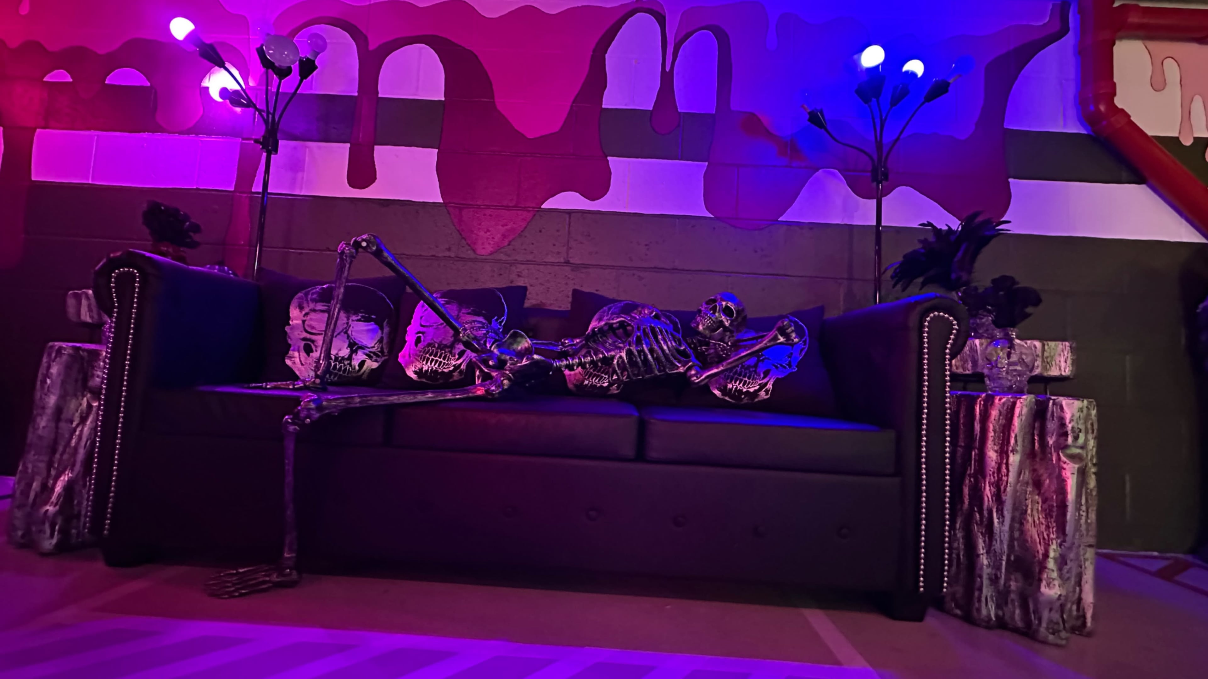 A skeleton rests on a black couch surrounded by colorful lights and patterned wall art.