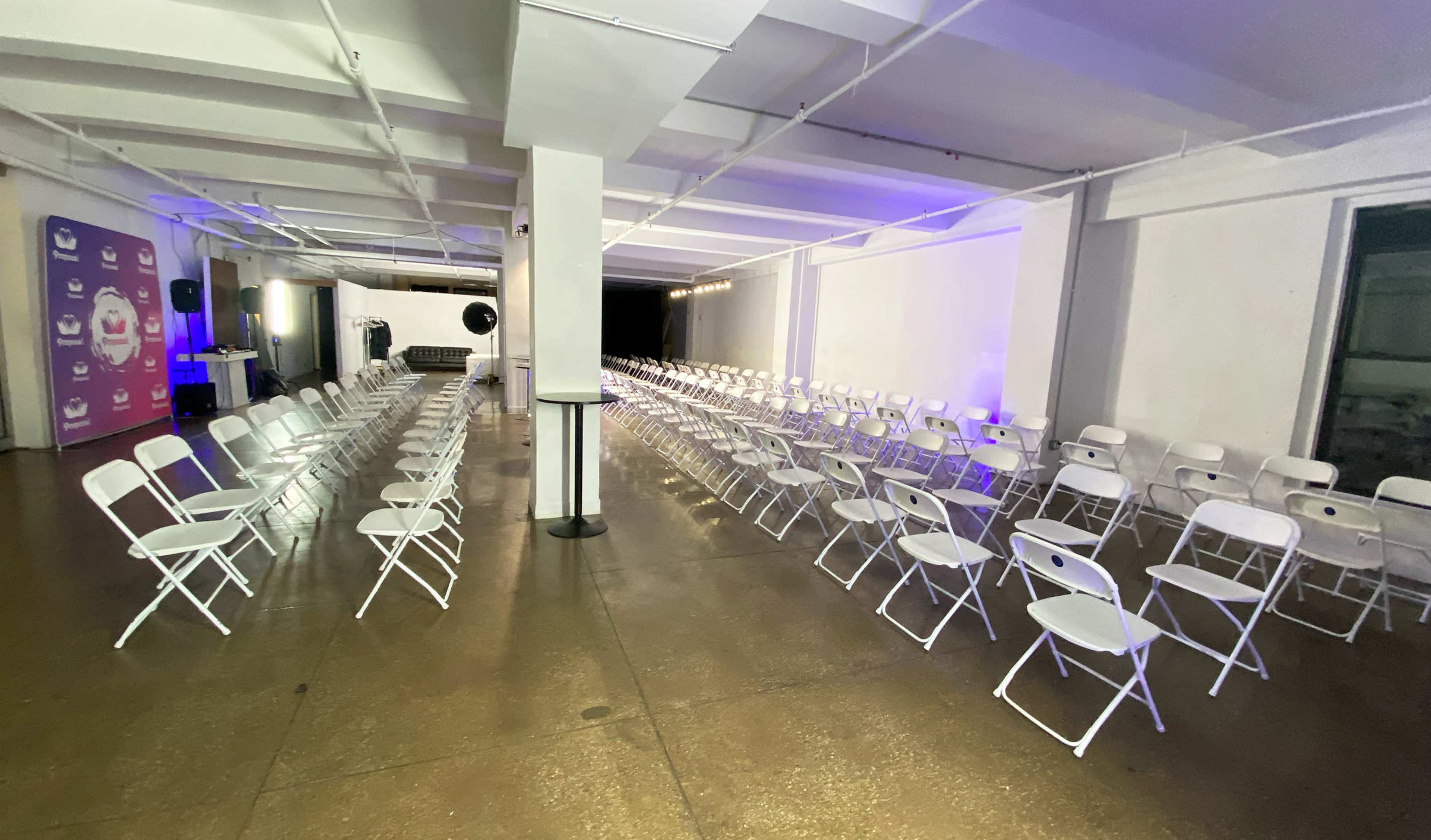 The image shows a spacious room with rows of white folding chairs arranged facing a stage area, and soft lighting illuminating the space.