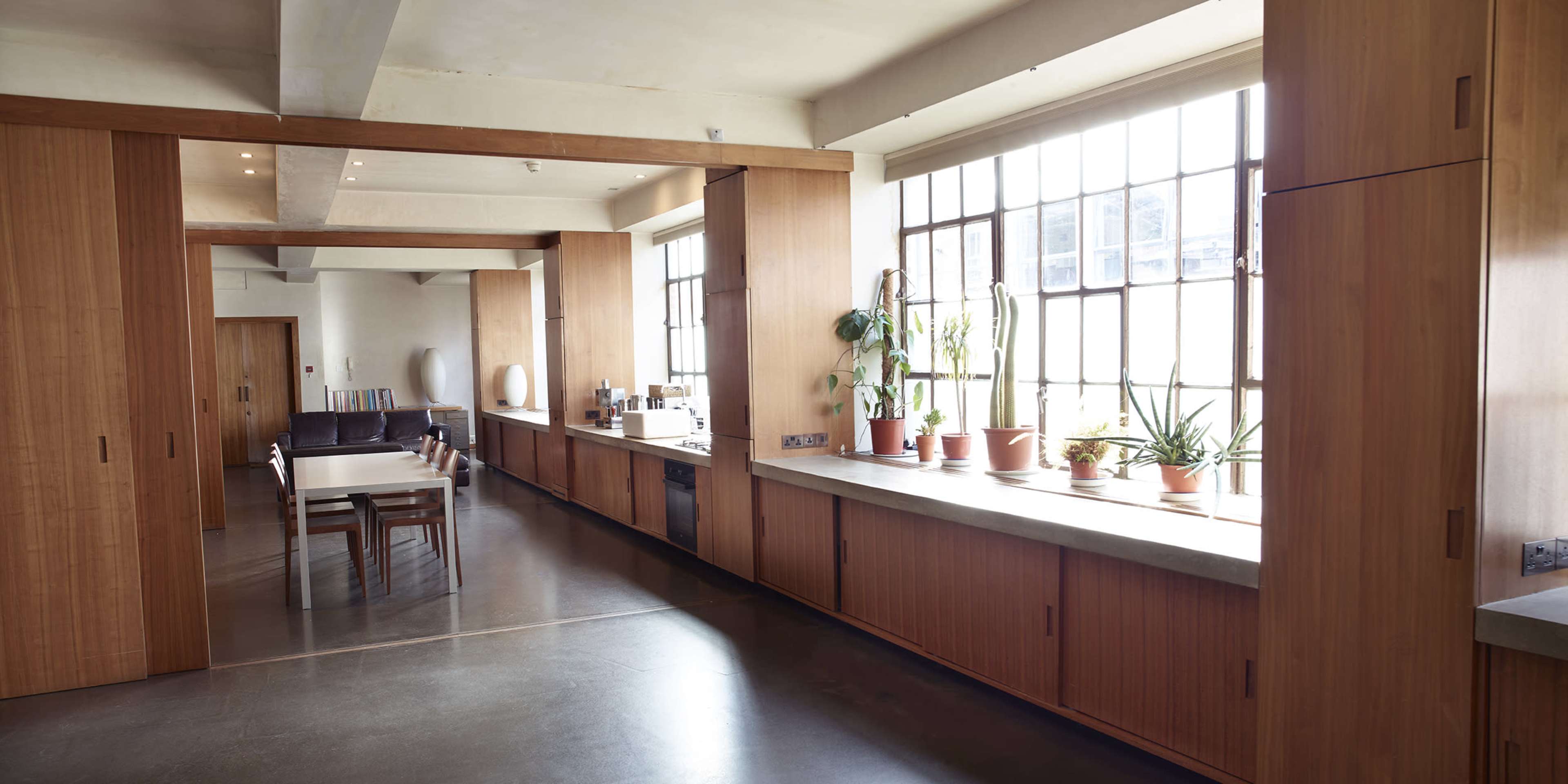 The image shows a modern kitchen and dining area with large windows, wooden cabinetry, and potted plants lining the counter.