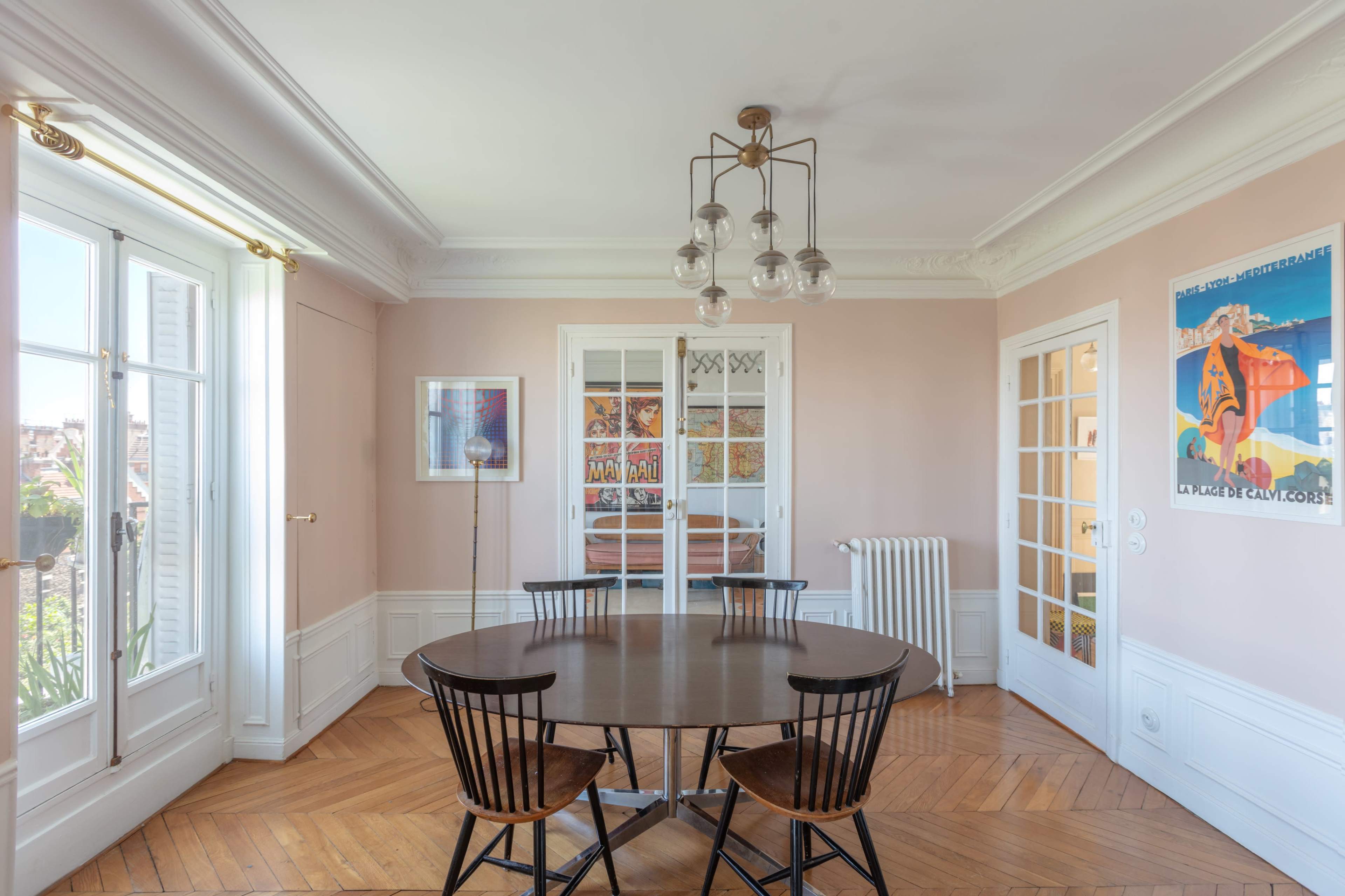The image shows a well-lit dining room featuring a round table surrounded by four black chairs, with a radiator and large windows on the walls.