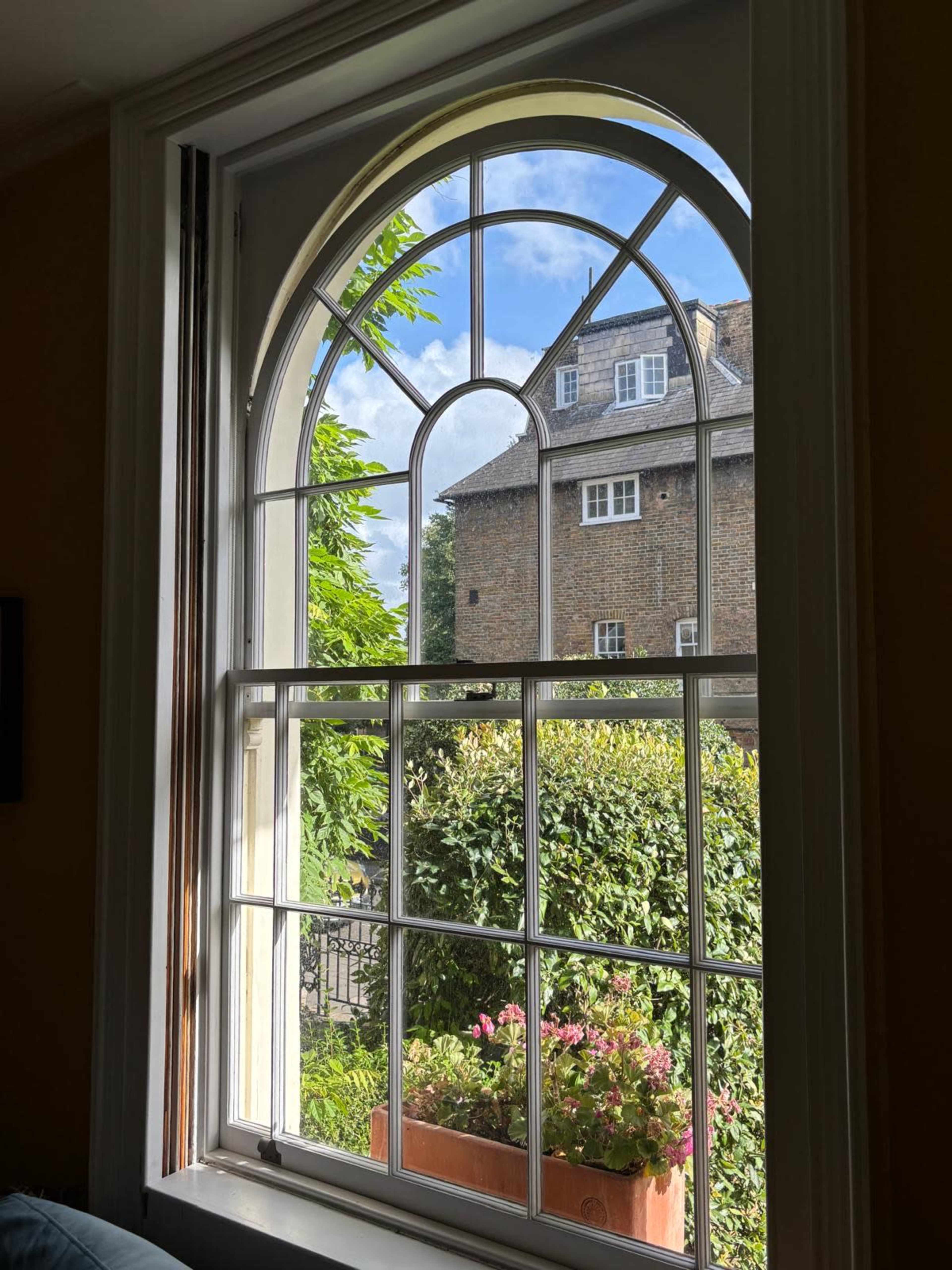 The image shows a large arched window with white frames, looking out onto a garden and a brick building under a partly cloudy sky.