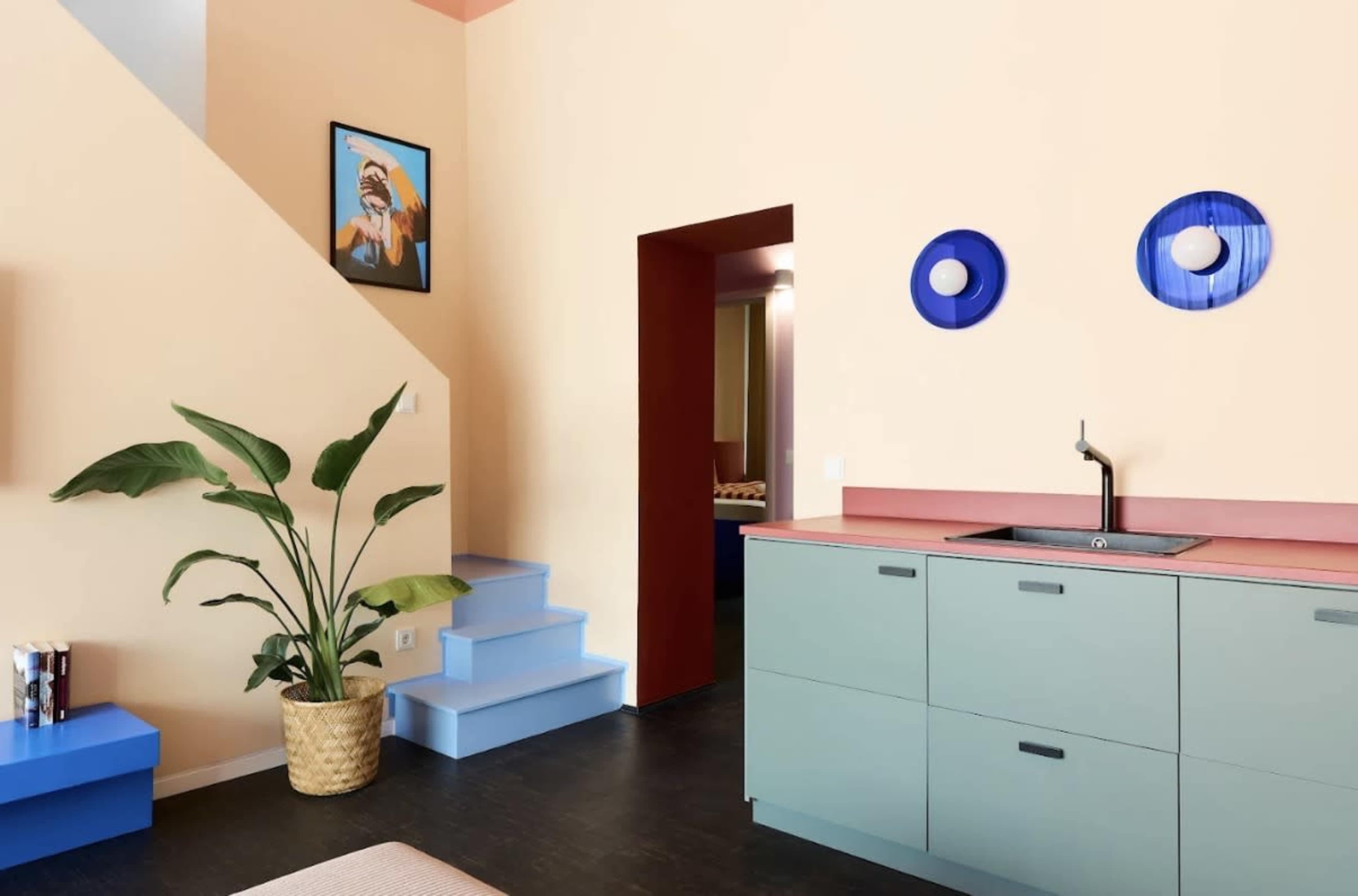 The image shows a brightly colored interior with a kitchen featuring green cabinets, a pink countertop, and blue wall decorations, alongside a staircase and a large potted plant.