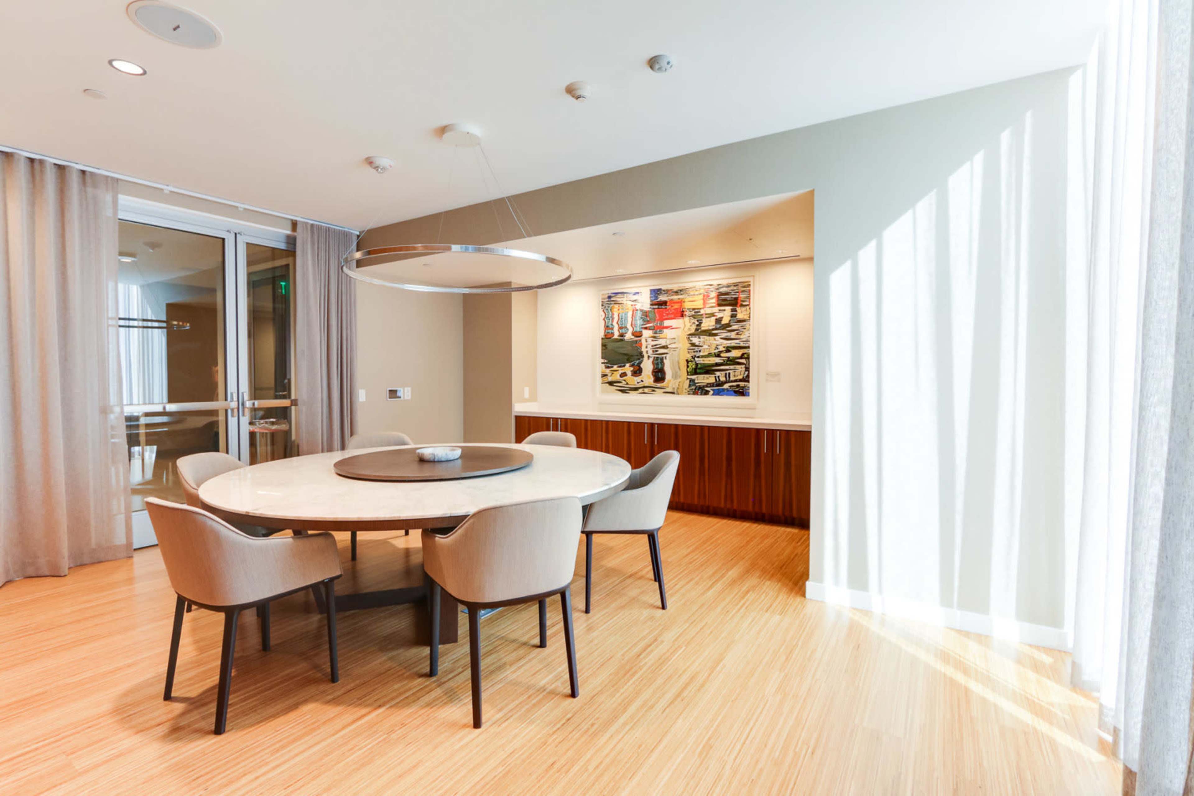 A round dining table with light-colored chairs is set in a brightly lit room featuring a large artwork on the wall.
