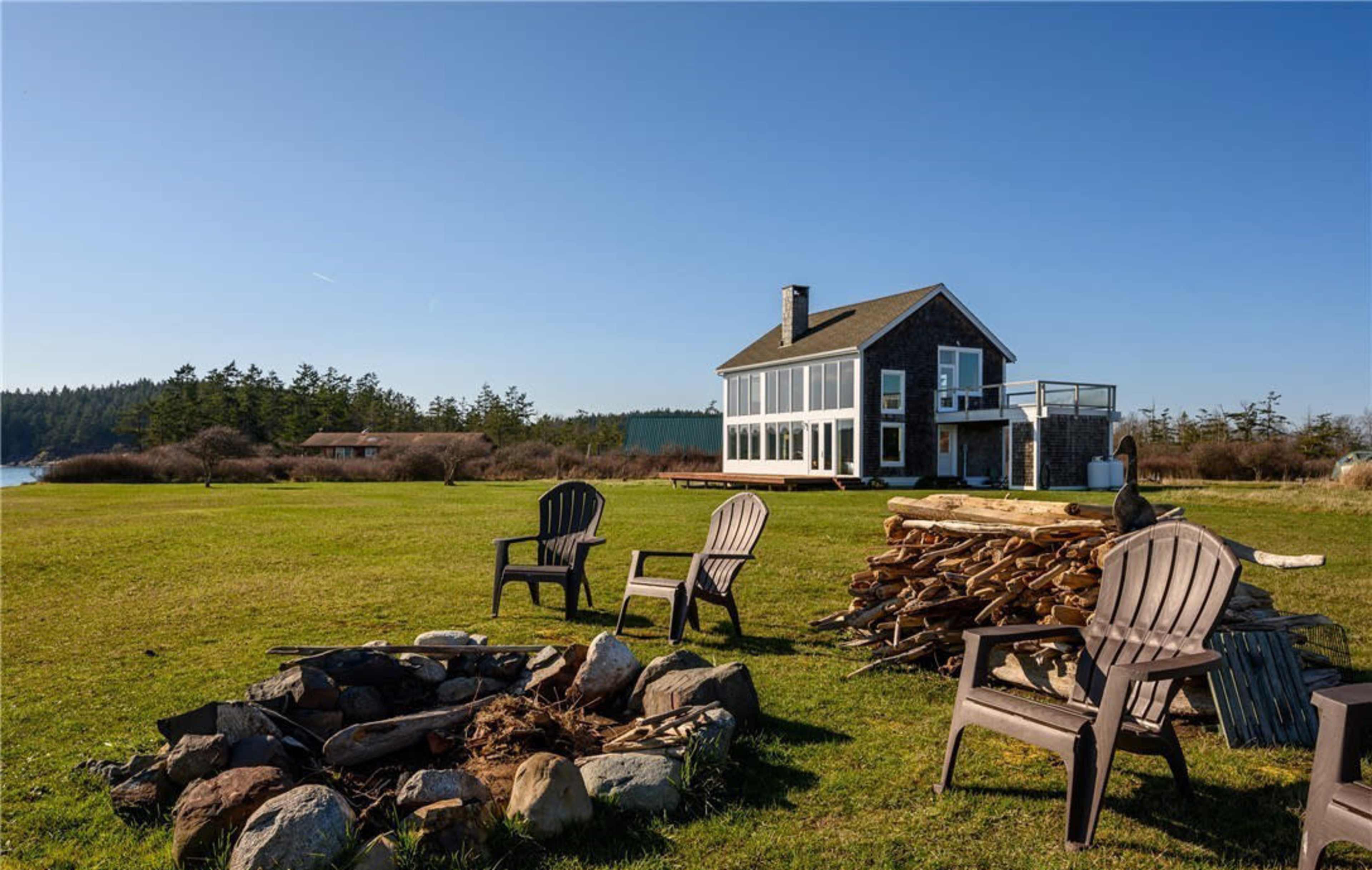 A house with large windows overlooks a grassy area with a fire pit surrounded by adirondack chairs and stacked firewood.
