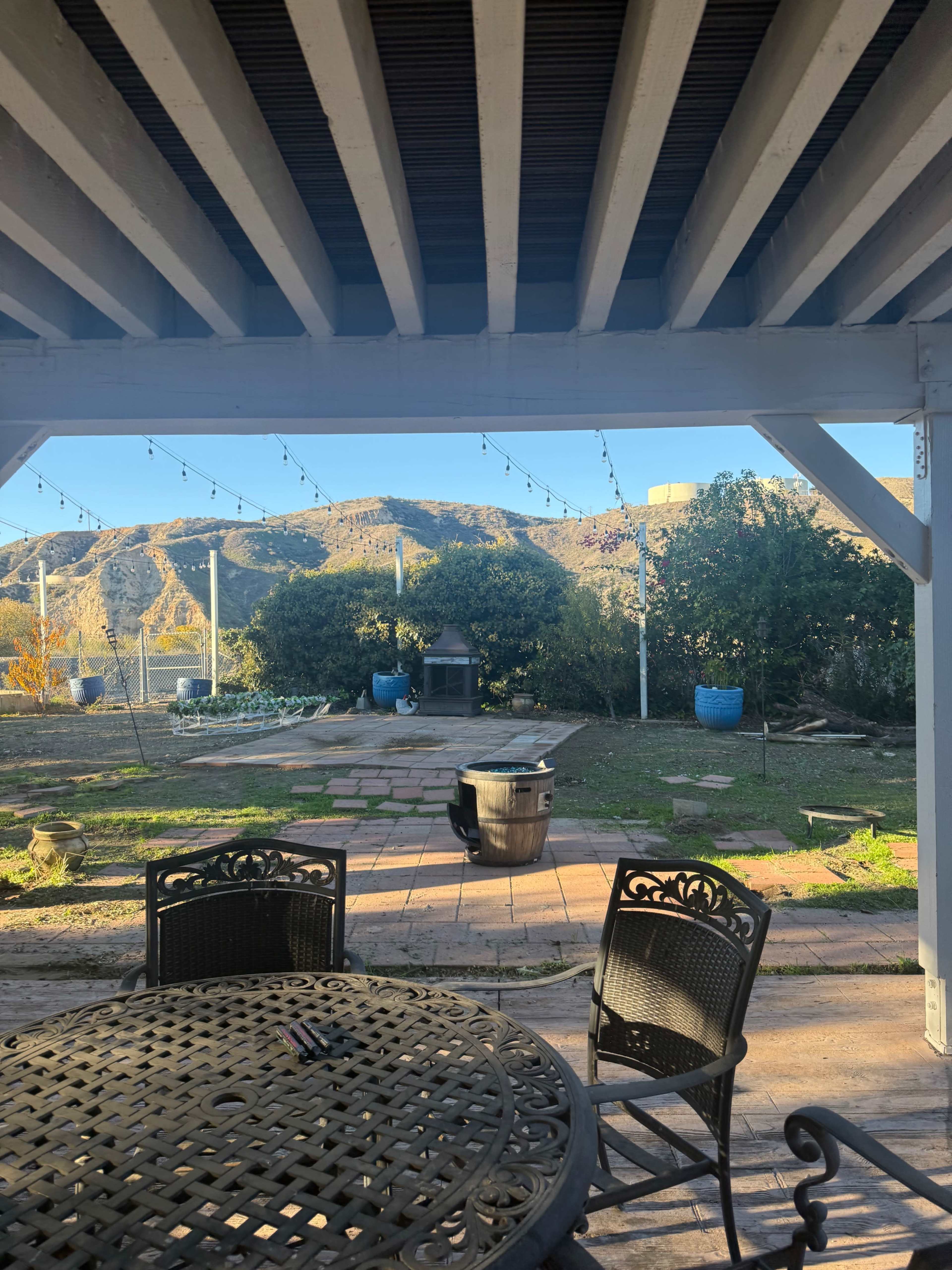The image shows a patio area with a round table and chairs, framed by a pergola, overlooking a grassy yard with a mountain backdrop and light strings overhead.