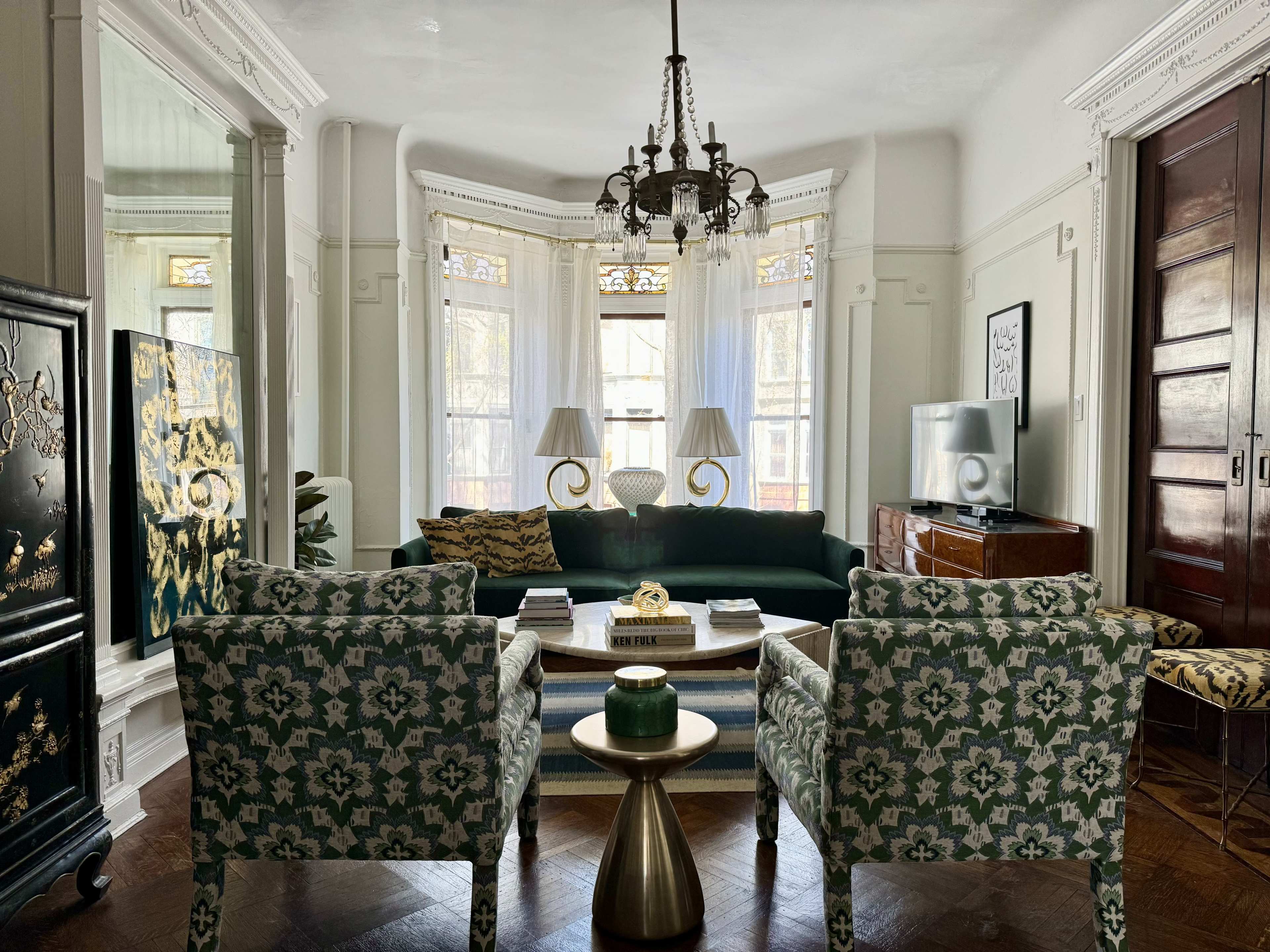 A brightly lit living room with a green sofa, two patterned armchairs, a coffee table, and large windows adorned with white drapes.