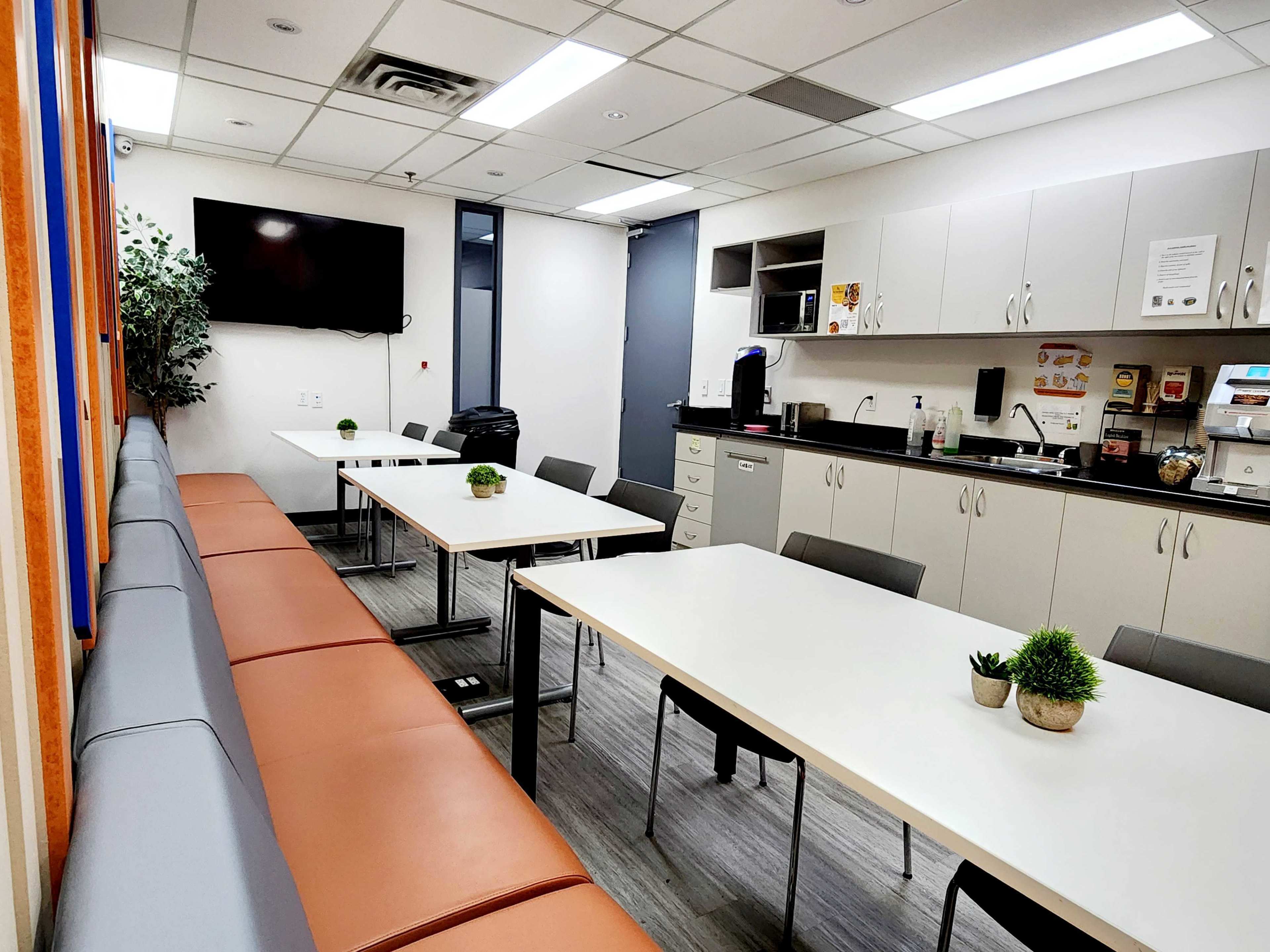 The image shows a modern break room with multiple tables, chairs, a kitchenette, and a television mounted on the wall.