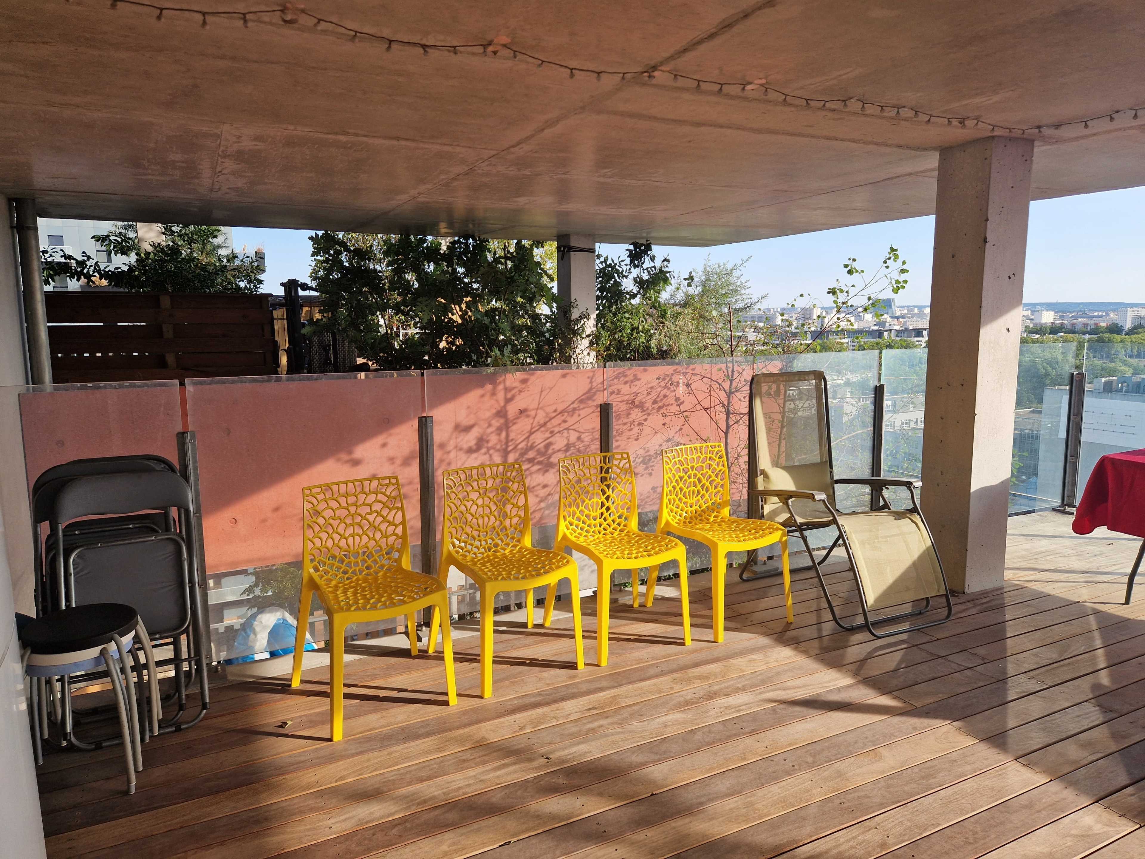 The image shows a covered outdoor patio with wooden flooring, featuring five yellow chairs arranged in a row and two other chairs positioned nearby.