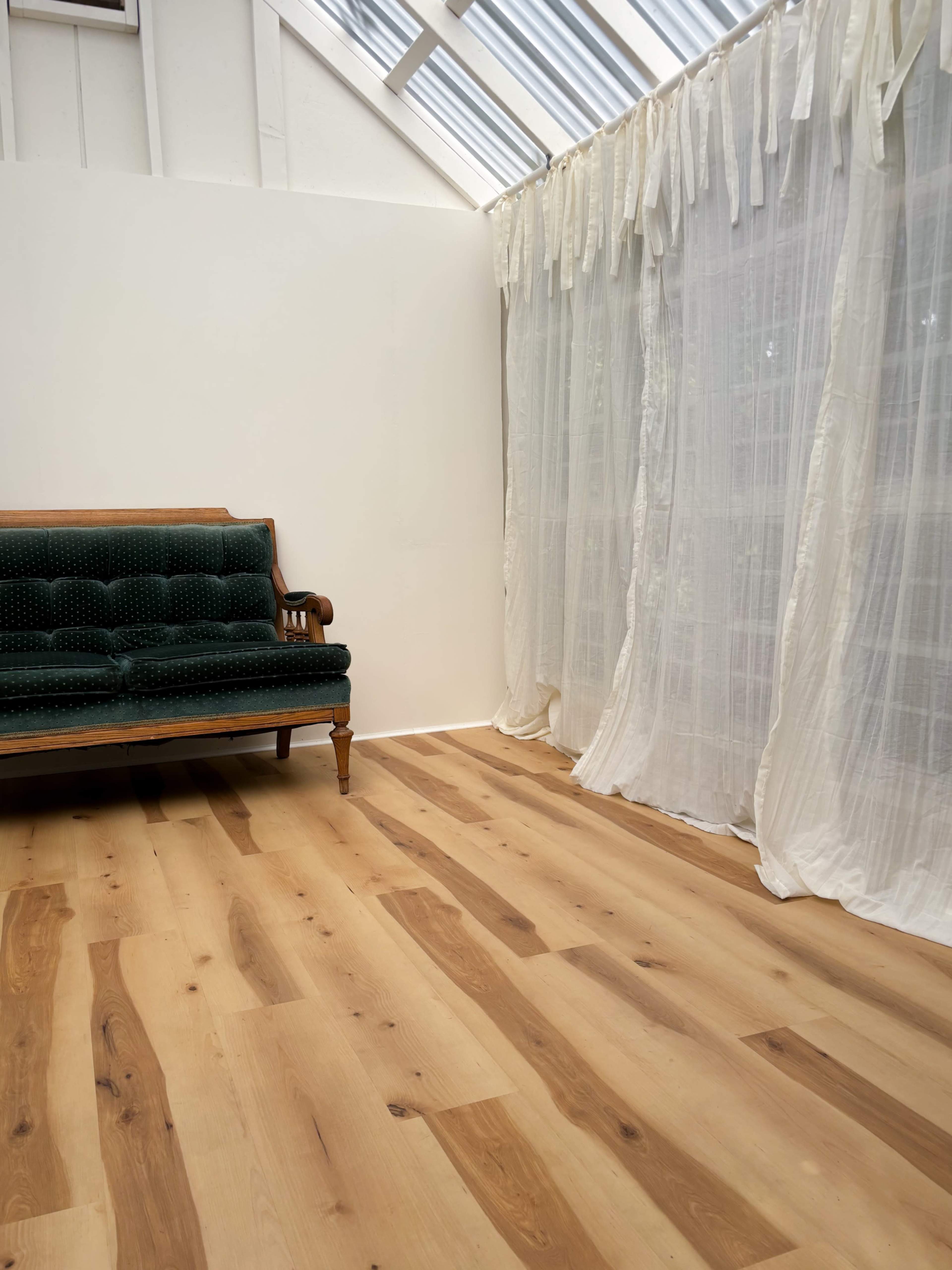 The image shows a minimalist interior with a green upholstered couch against a white wall, next to sheer white curtains and light wood flooring.