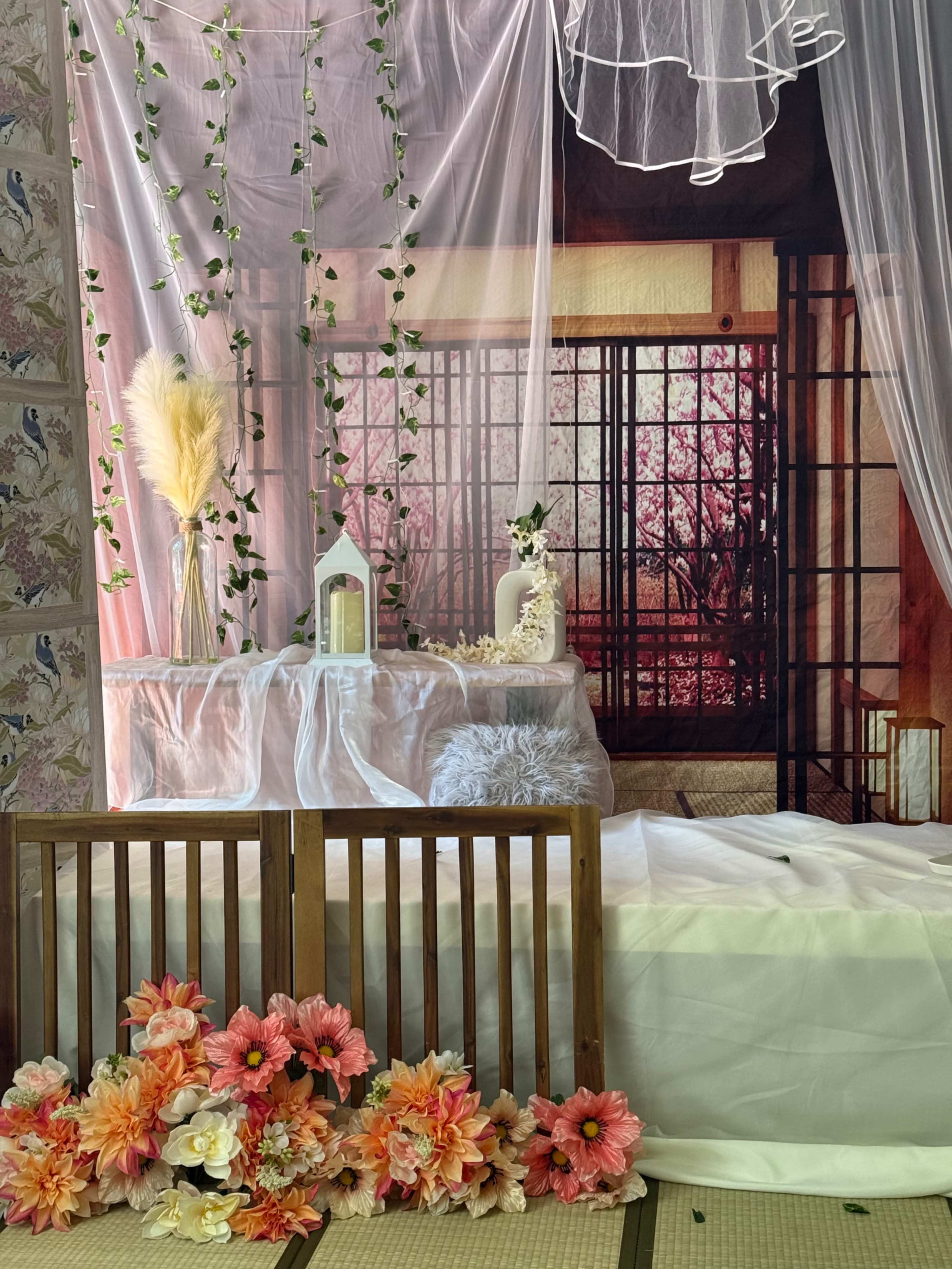 The image shows a decorative room with a floral arrangement, soft fabrics, and a backdrop featuring a traditional window with a view of pink flowers.