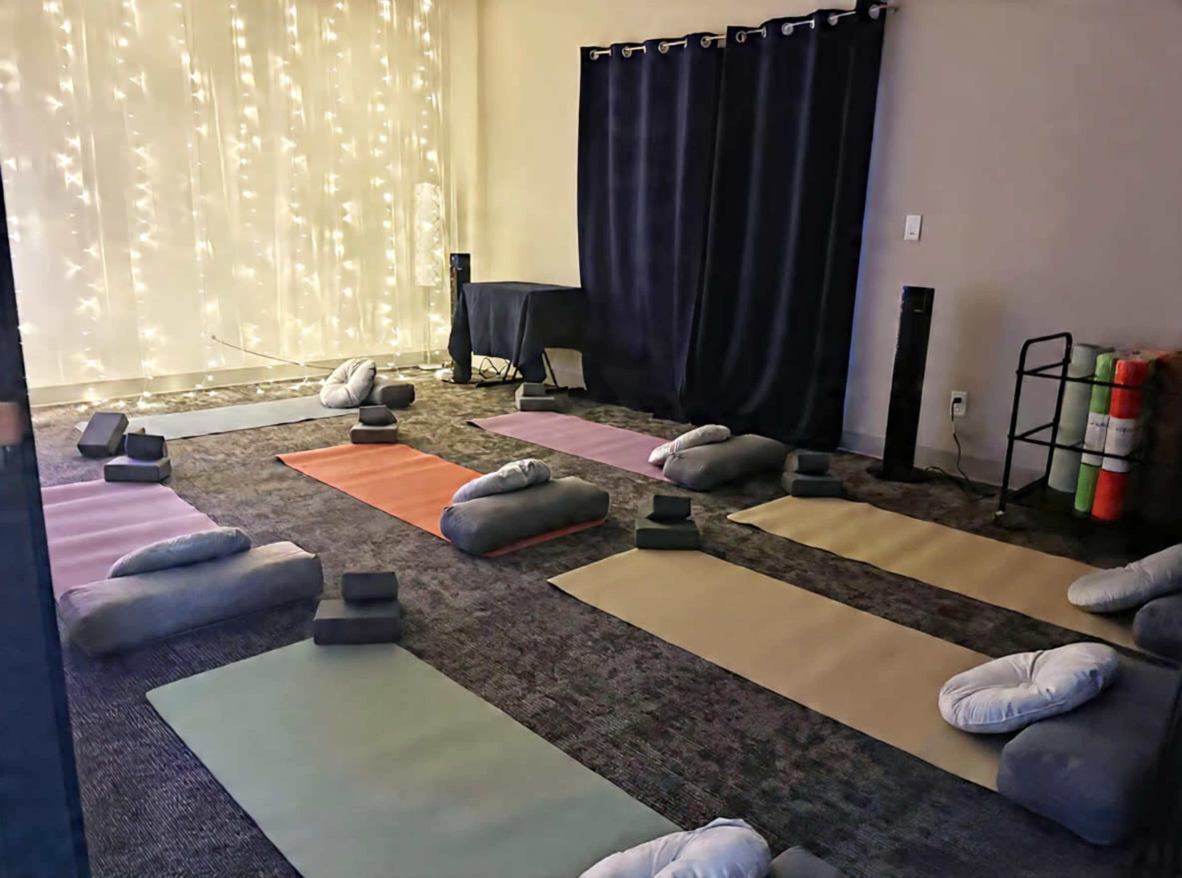 The image shows a yoga studio with several yoga mats arranged on the floor, cushions, and blocks, illuminated by soft lights in the background.