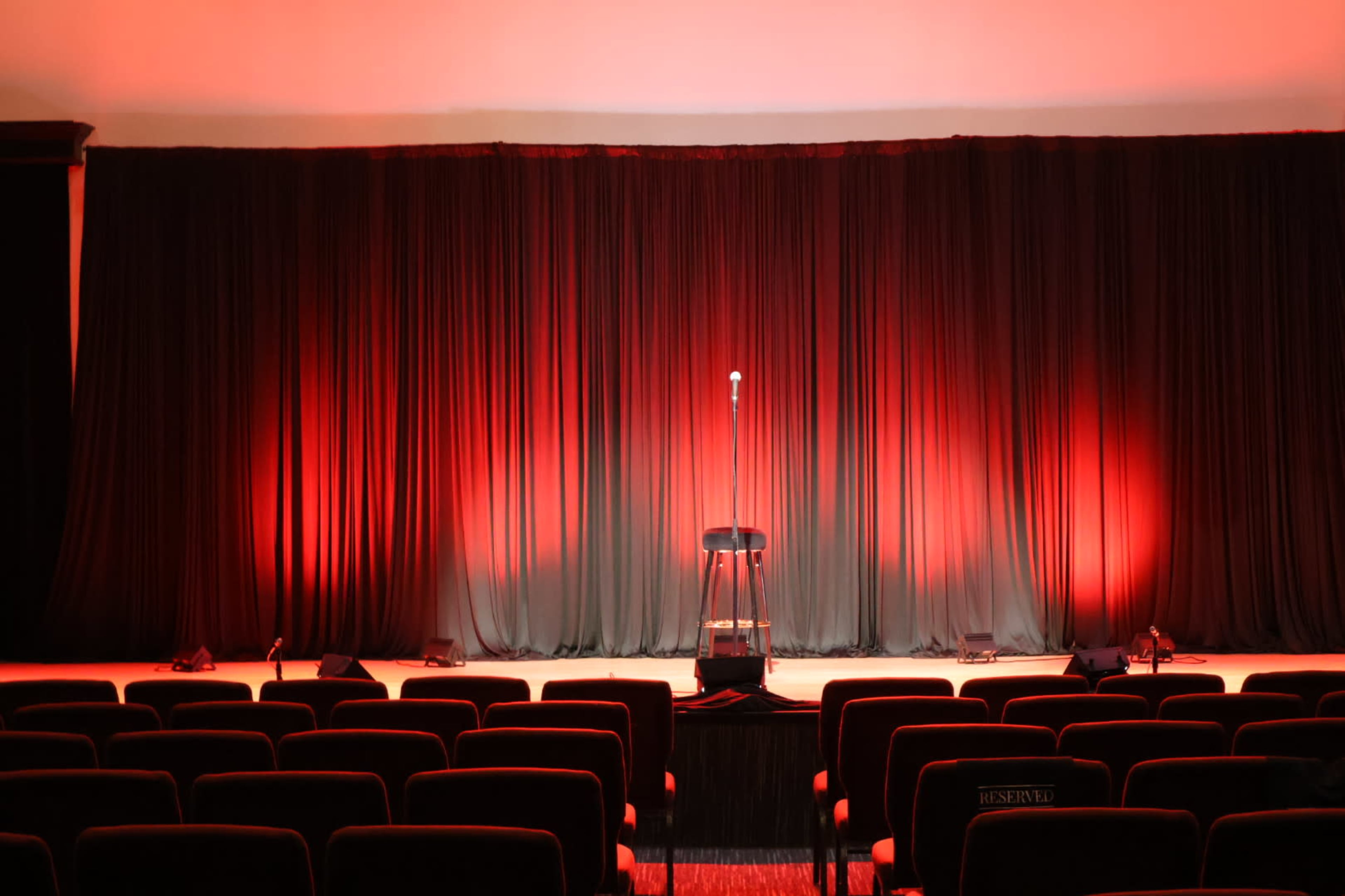 A spotlight shines on a microphone stand and stool in front of a dark curtain, with red lighting illuminating the stage.
