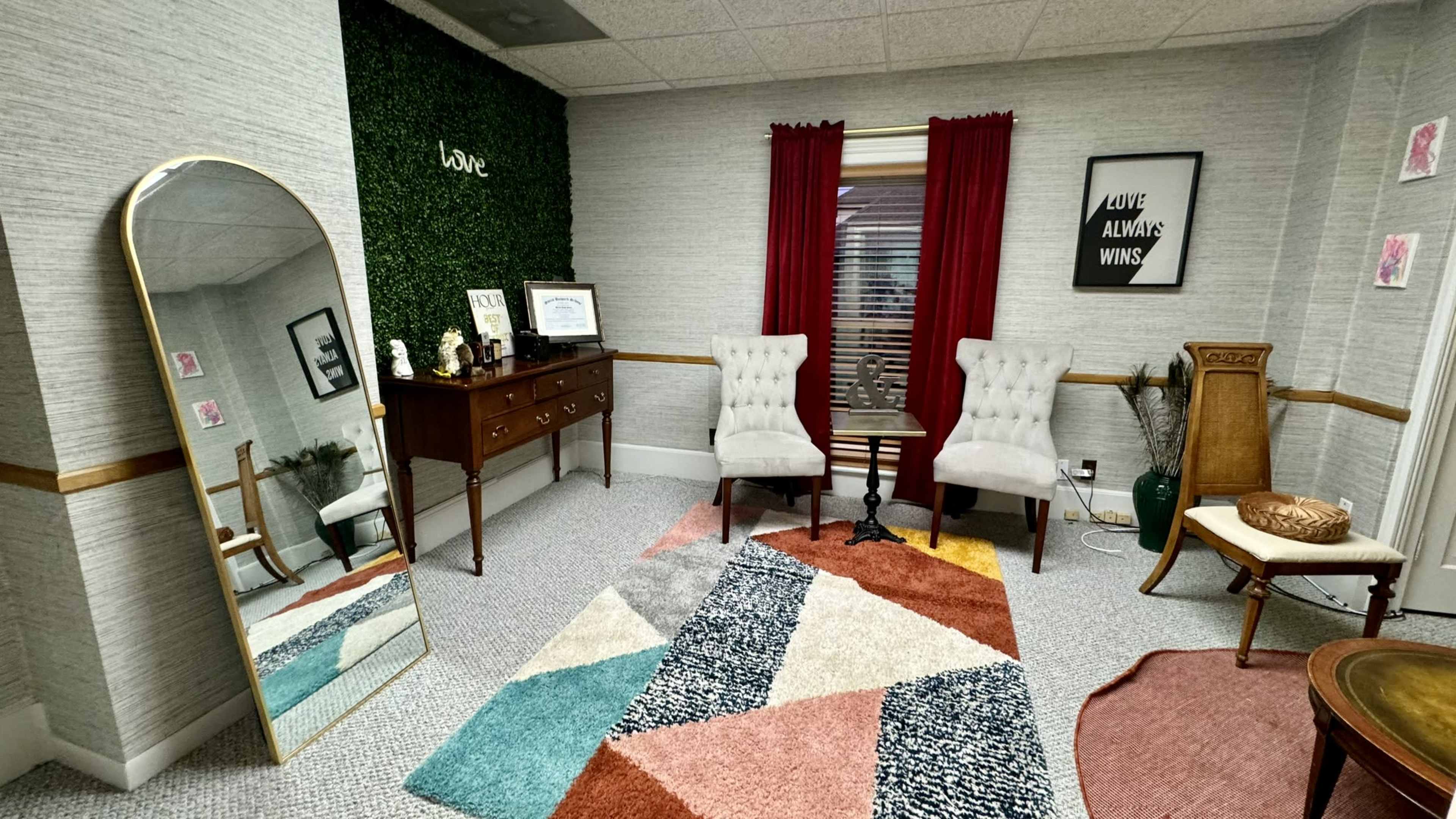 The room features a large mirror, two upholstered chairs, a wooden dresser, and a colorful geometric rug.