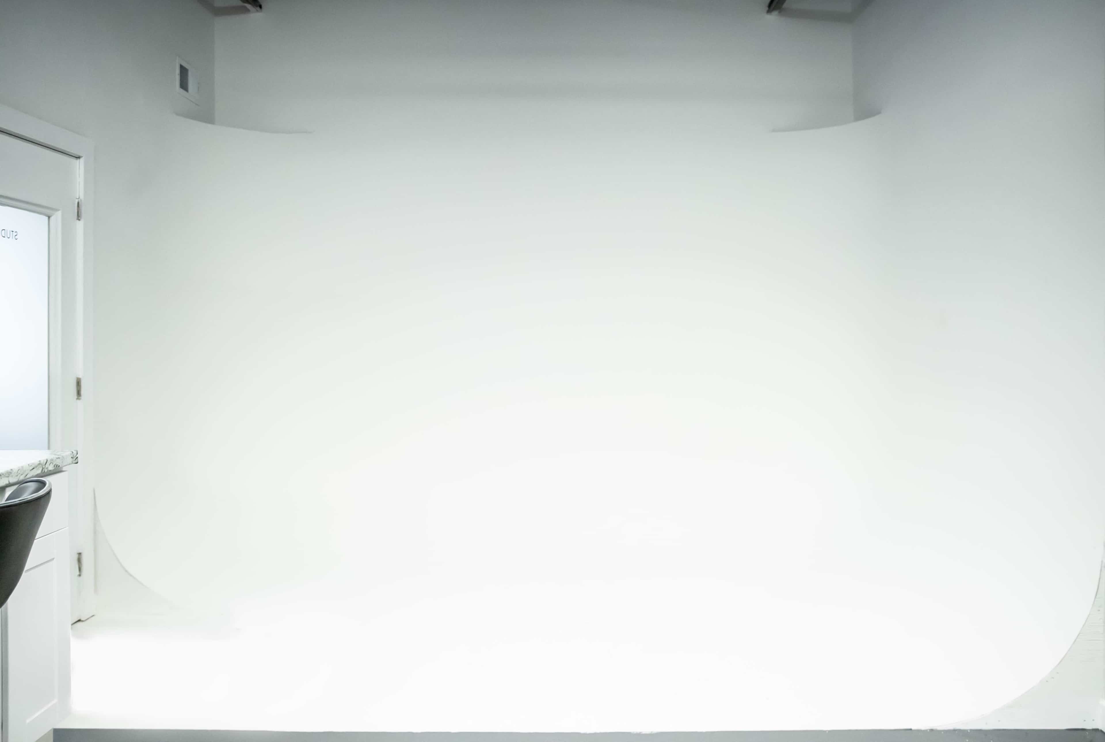 A brightly lit, minimalist studio space with a seamless white backdrop and a door on the left side.