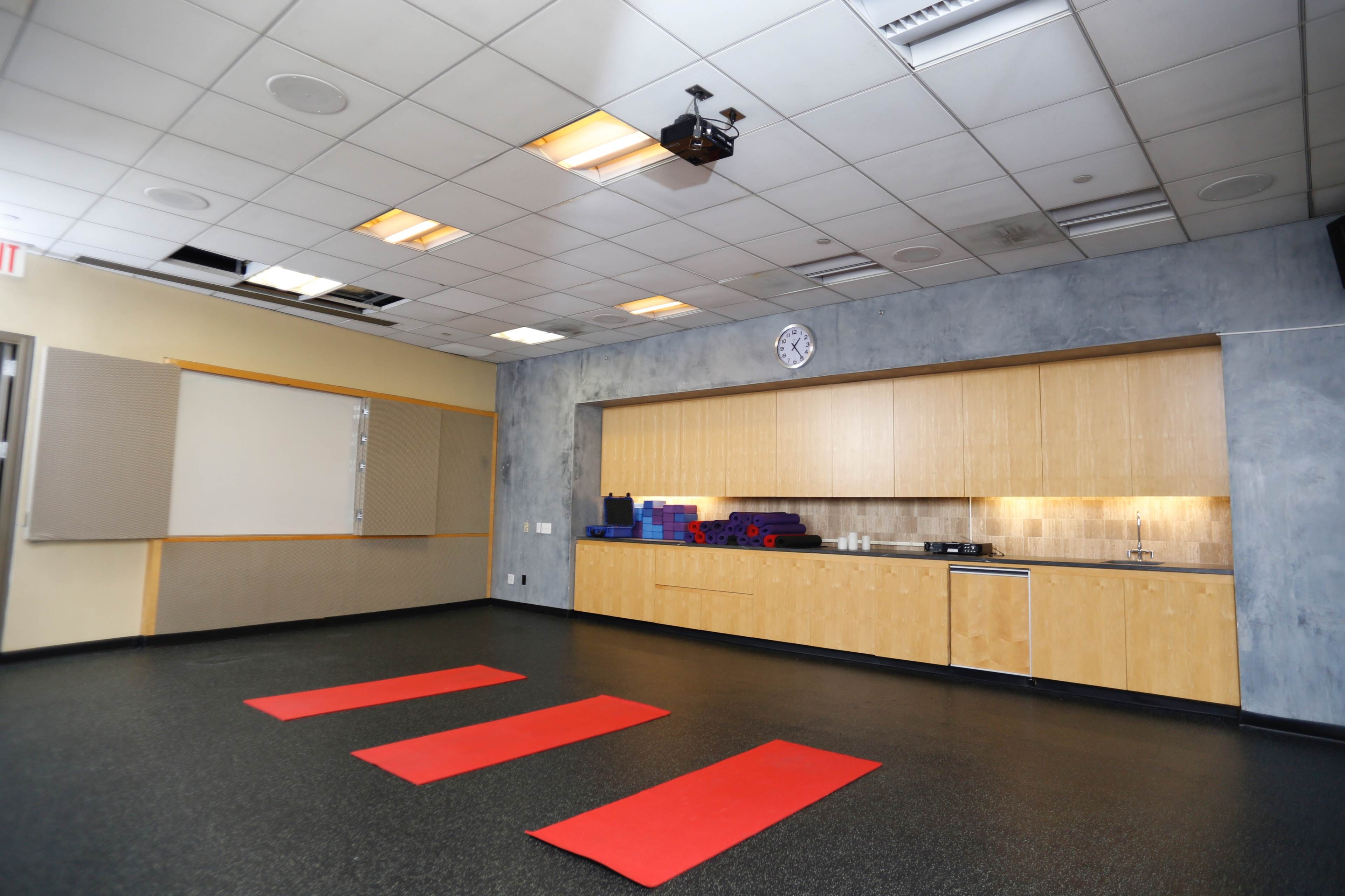 The image shows a yoga studio with three red mats on the floor, a kitchen area with wooden cabinets, and a clock mounted on the wall.