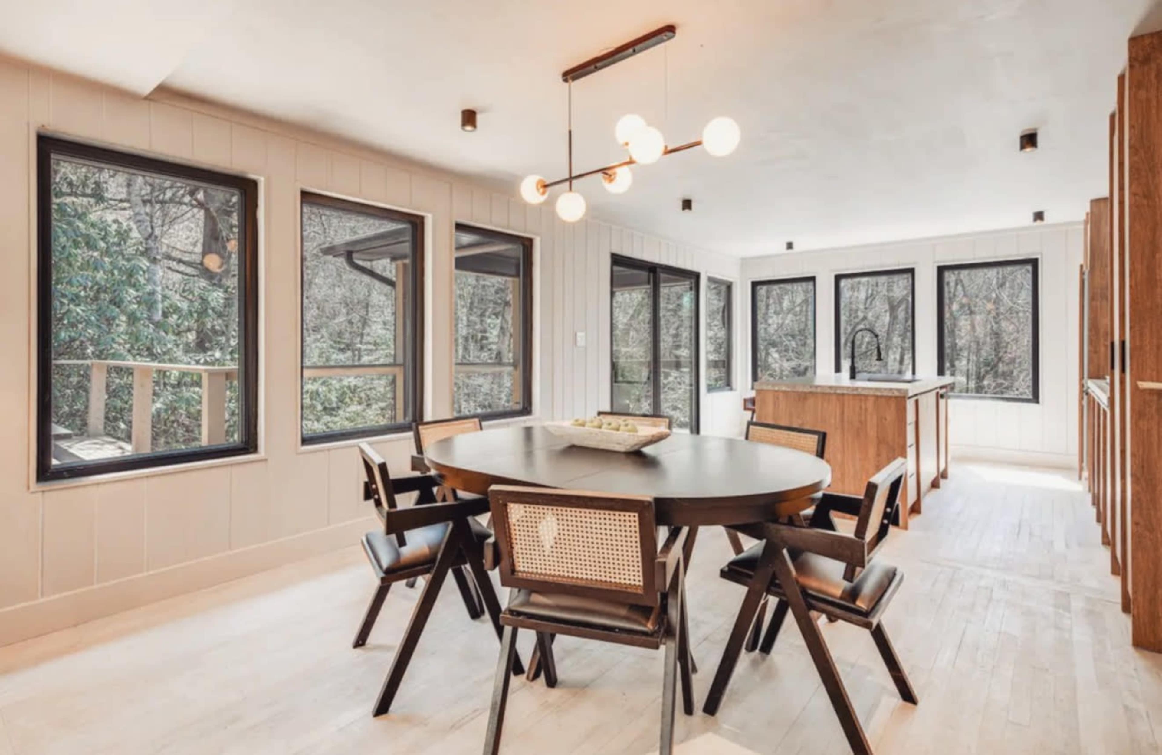 The image shows a bright, modern dining area featuring a circular table surrounded by black chairs, large windows that offer a view of the outdoors, and a kitchen area in the background.
