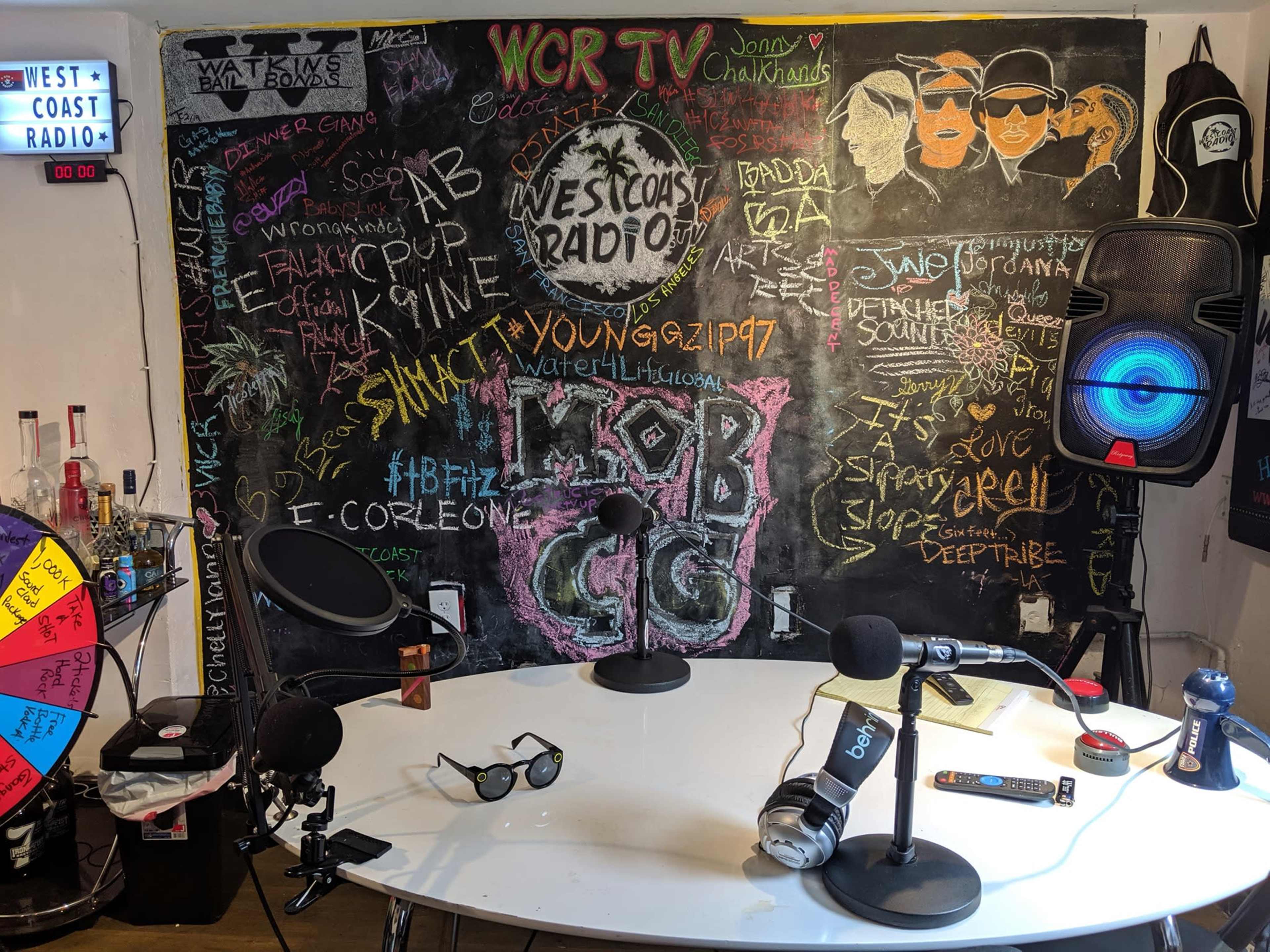 A radio studio with a circular table, microphones, a colorful chalkboard filled with messages, and a large speaker.