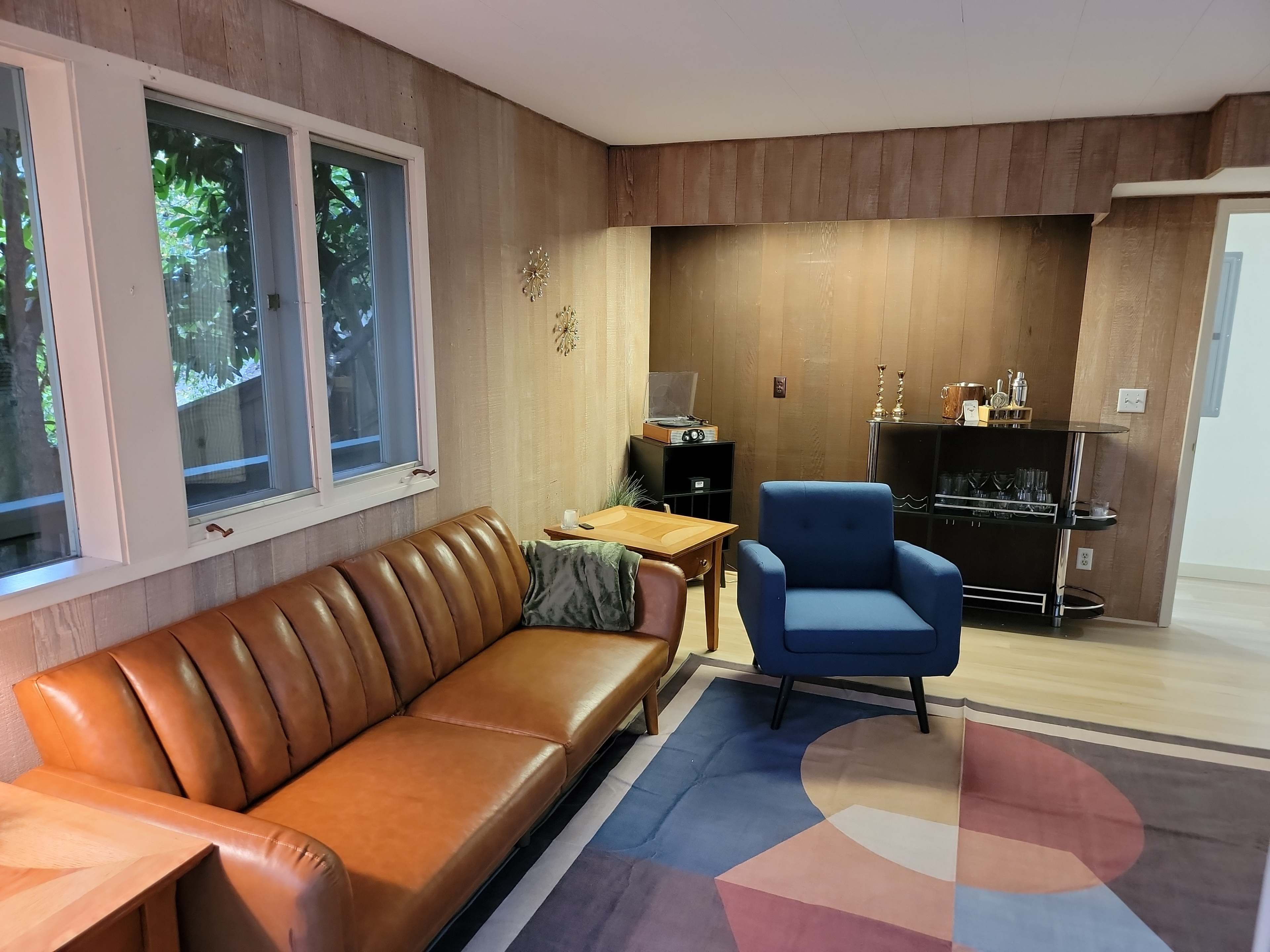 The living room features a brown leather sofa, a blue armchair, and a wooden bar cabinet against textured wood paneling.