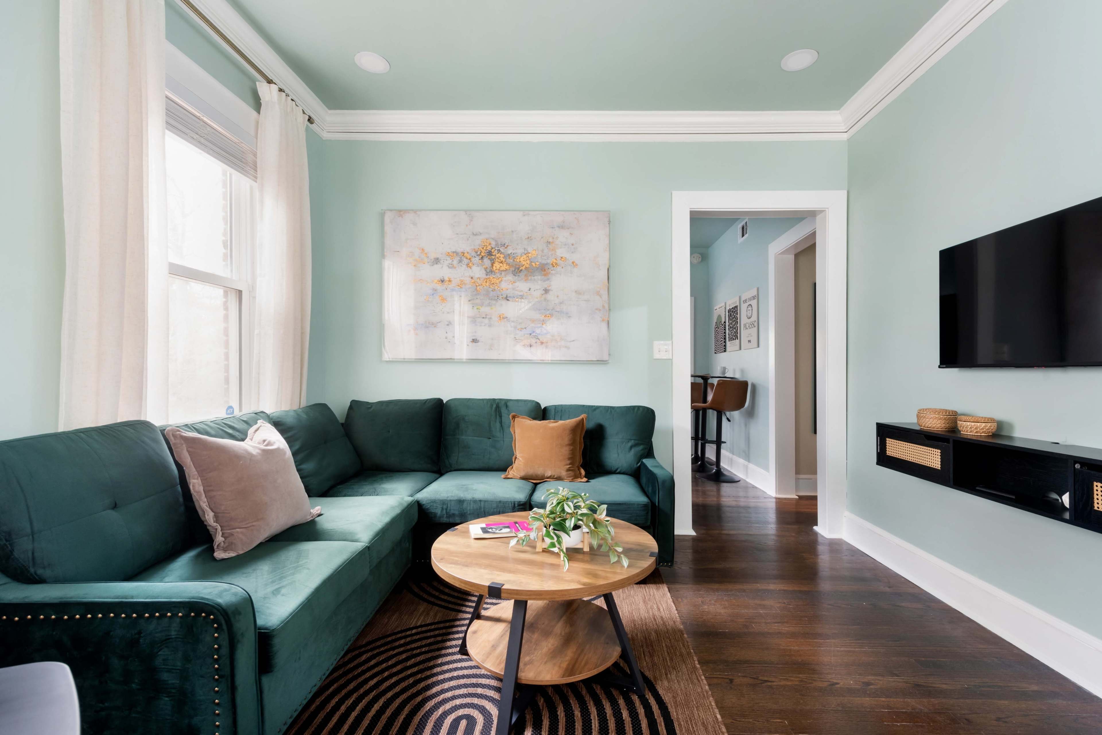 A cozy living room features a green sectional sofa, a round wooden coffee table, and light-colored walls with artwork hanging above.
