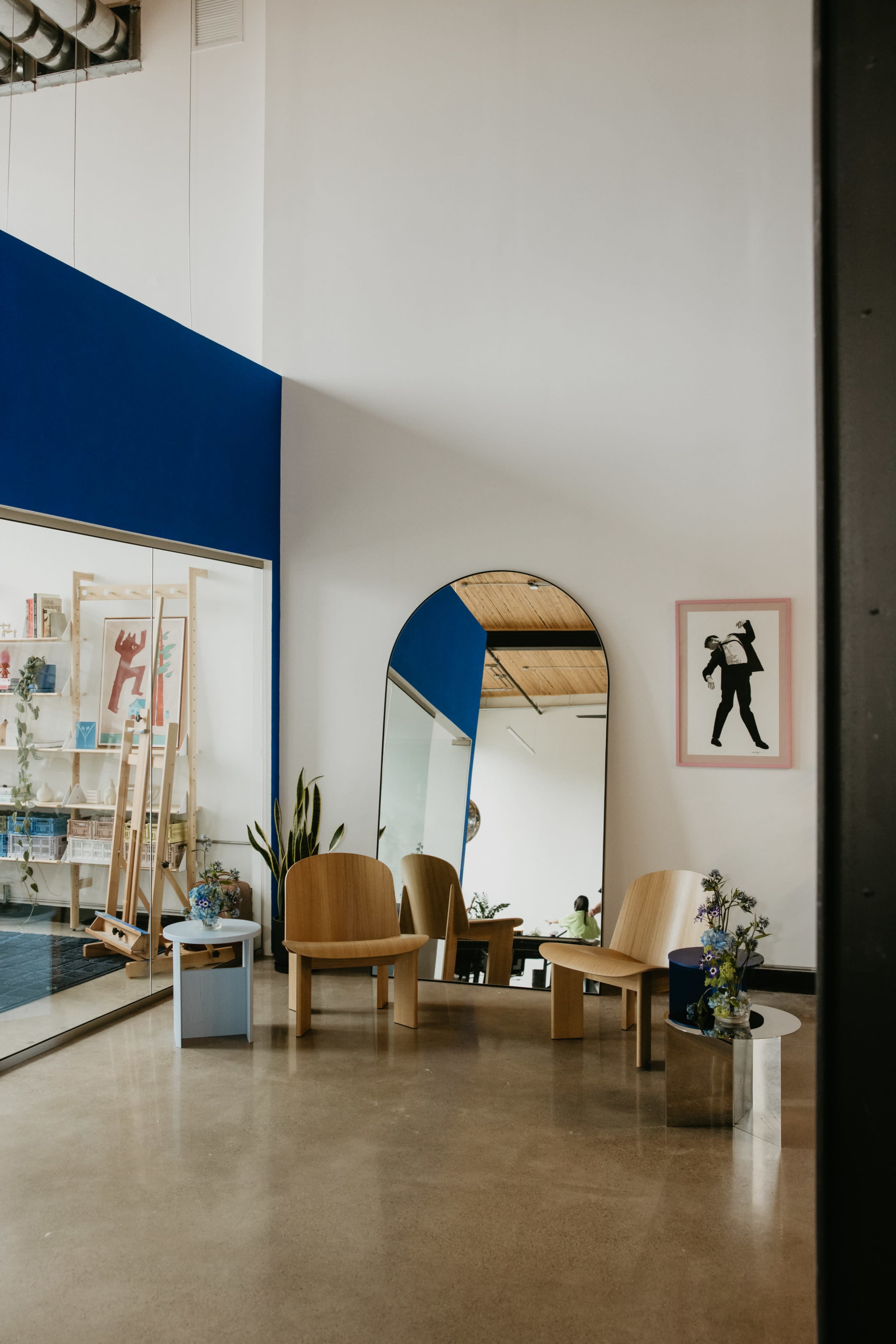 The image shows a modern interior space featuring two wooden chairs, a large mirror, and a vibrant blue accent wall with artwork.