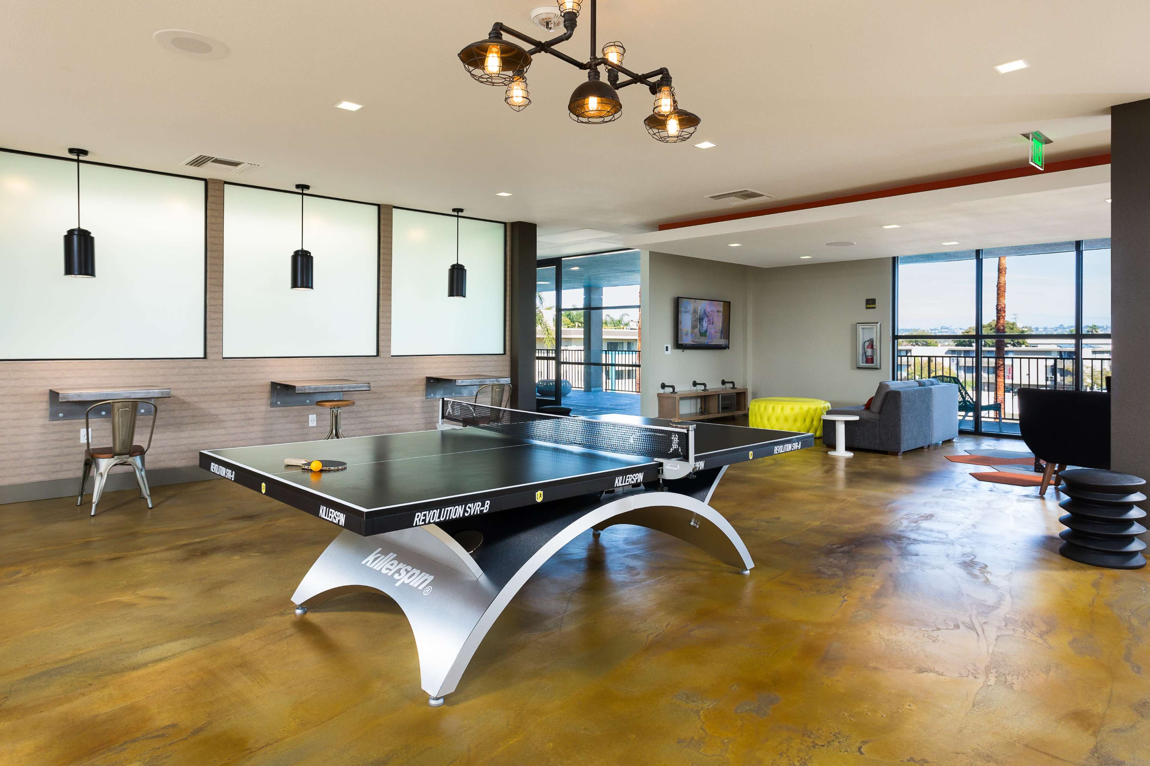The image shows a modern recreational room featuring a ping pong table, lounge seating, and large windows providing natural light.