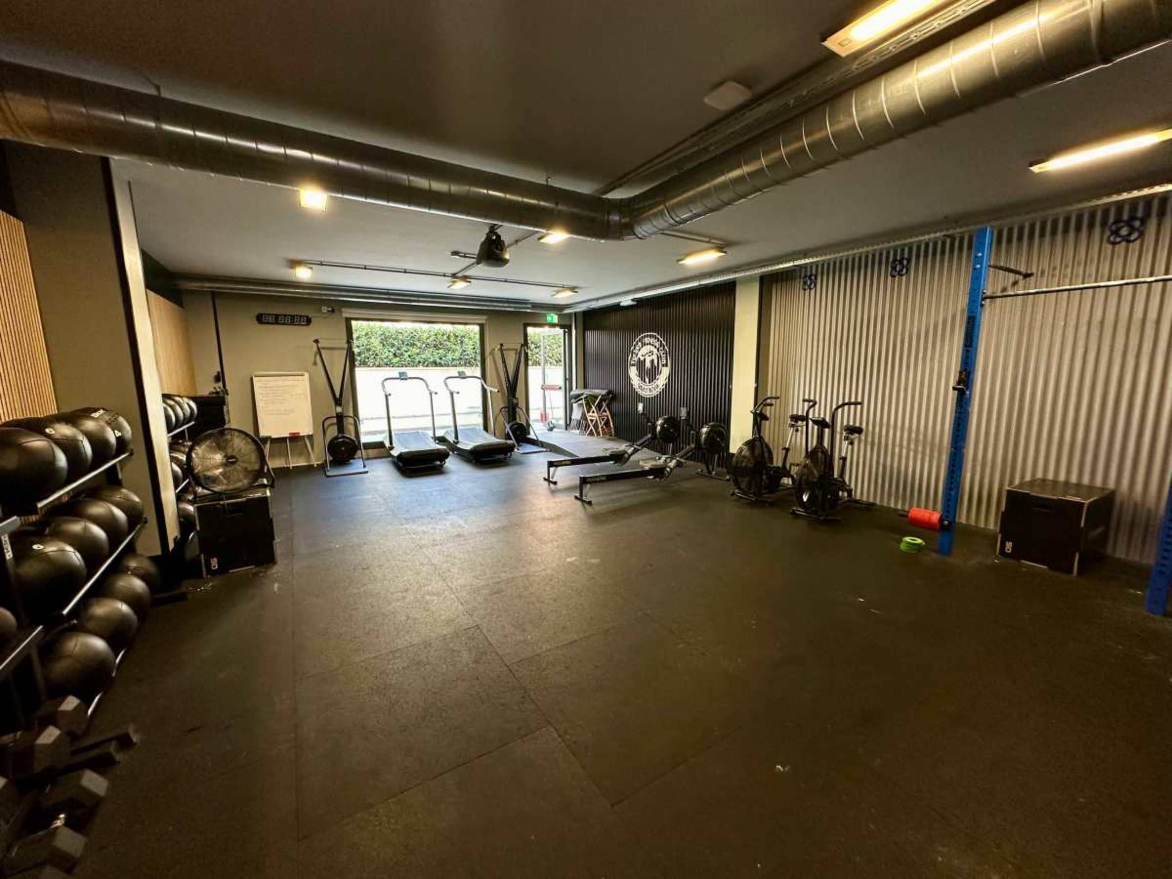 An interior gym space equipped with various exercise machines, weights, and mats, featuring large windows that allow natural light to enter.