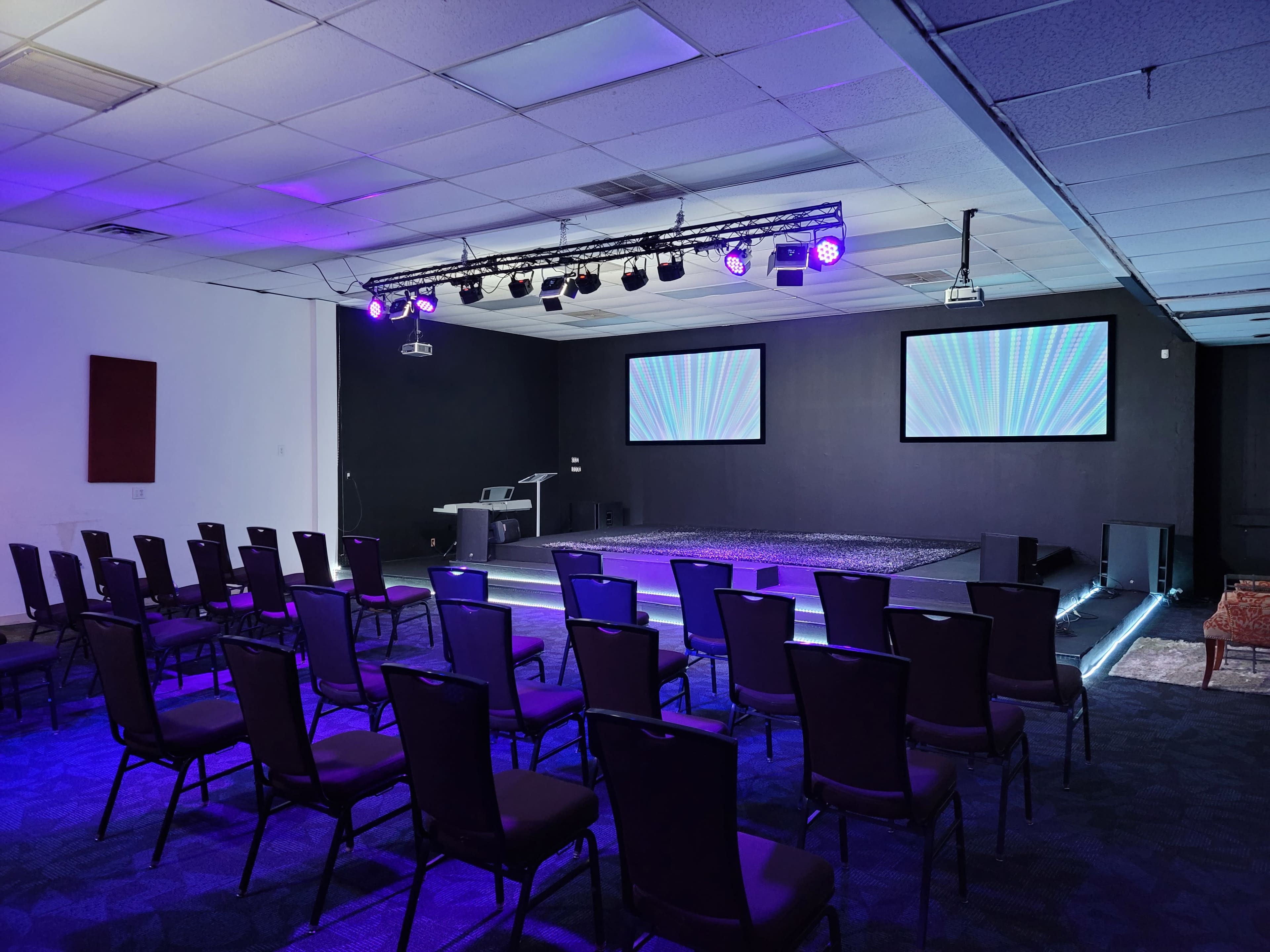 A prepared indoor event space with rows of chairs facing a stage that has two projection screens and stage lighting.