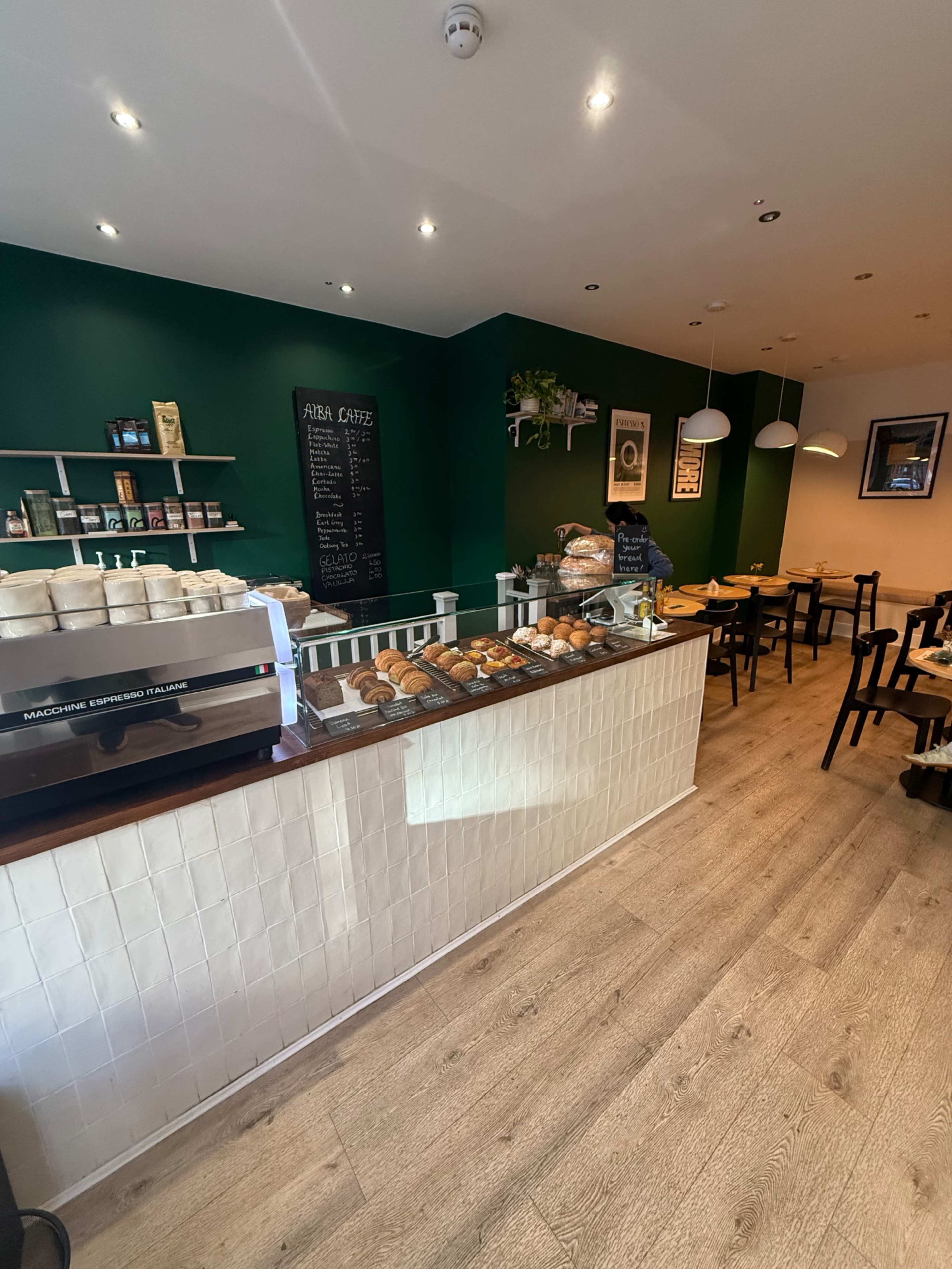 The image depicts the interior of a café with a brightly lit space featuring a counter displaying baked goods and green accent walls, along with wooden tables and chairs.