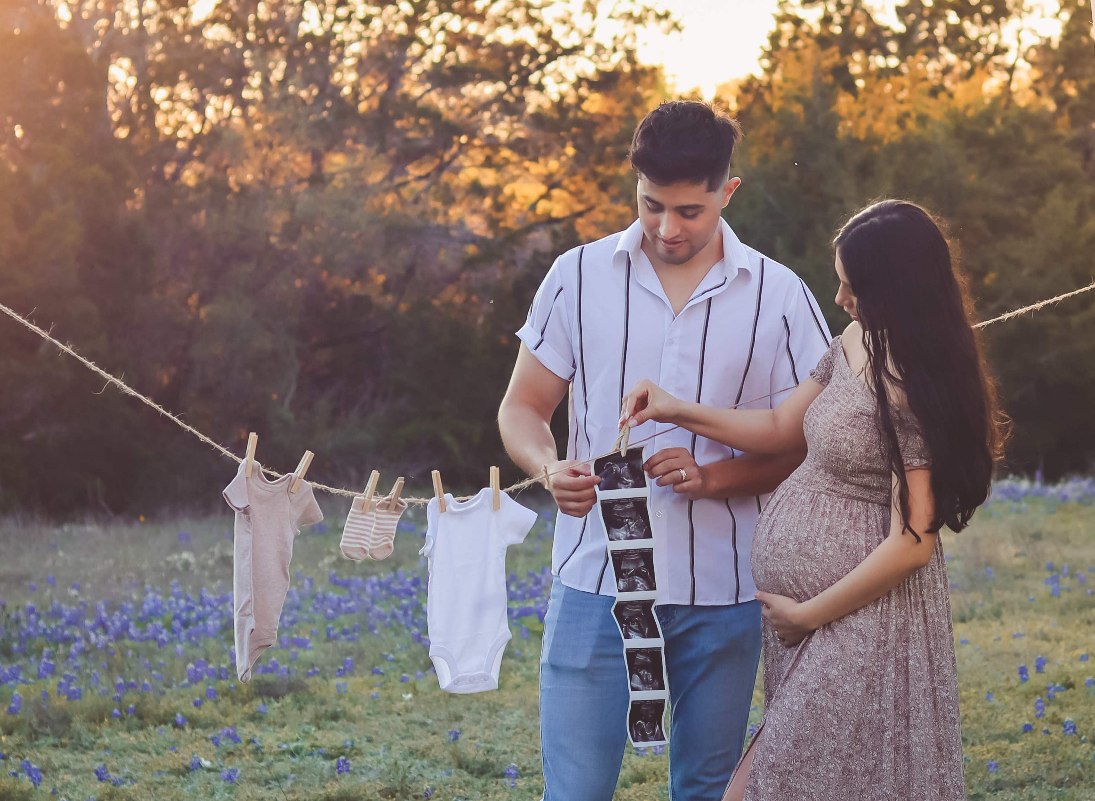 A couple stands in a field with blue flowers, holding ultrasound photos and hanging baby clothes on a line.