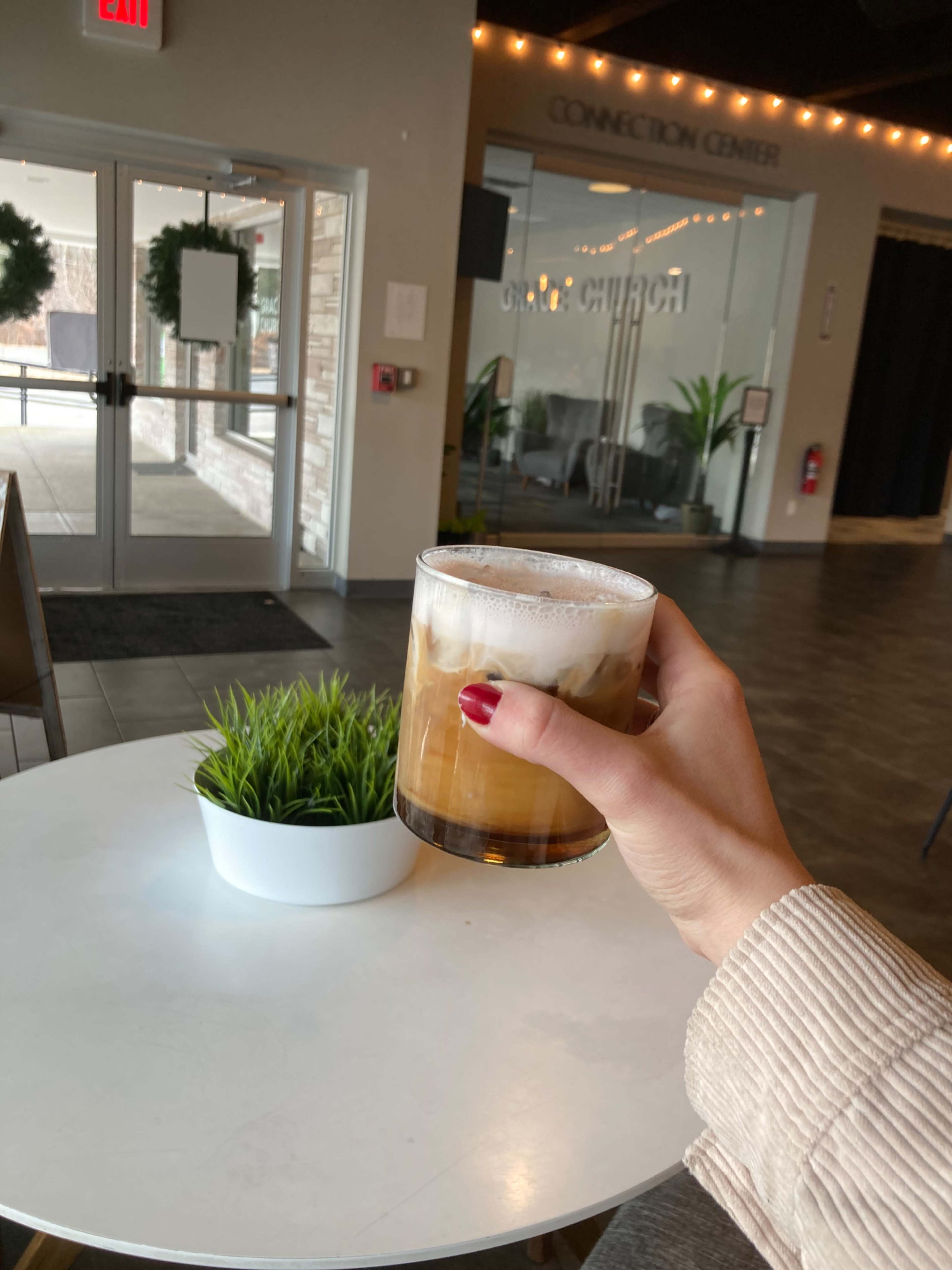 A person holds a glass of iced coffee in a modern interior space with a potted plant on a white table.