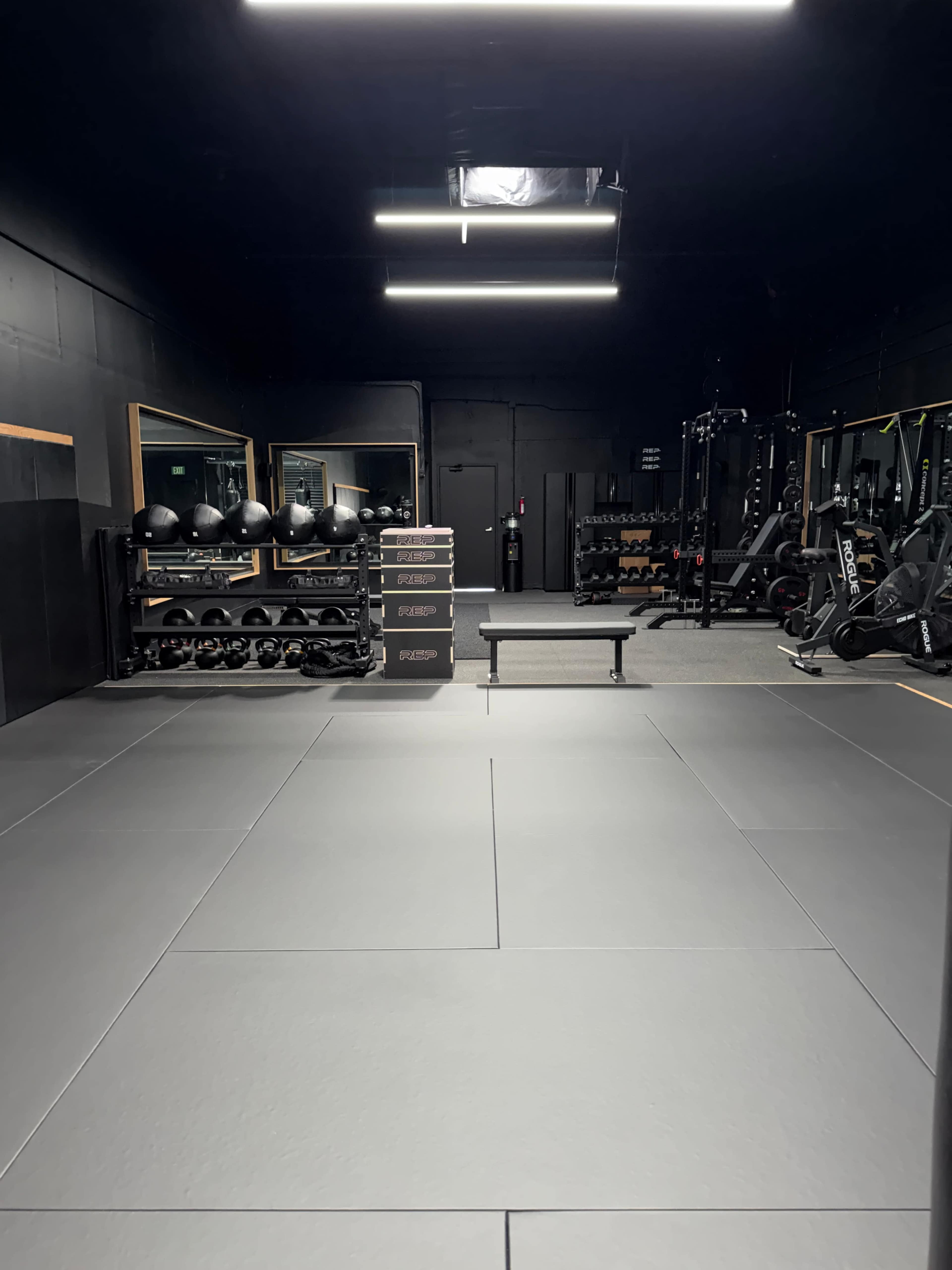 The image shows an empty gym interior with mats on the floor, a bench in the center, and various exercise equipment stored along the walls.