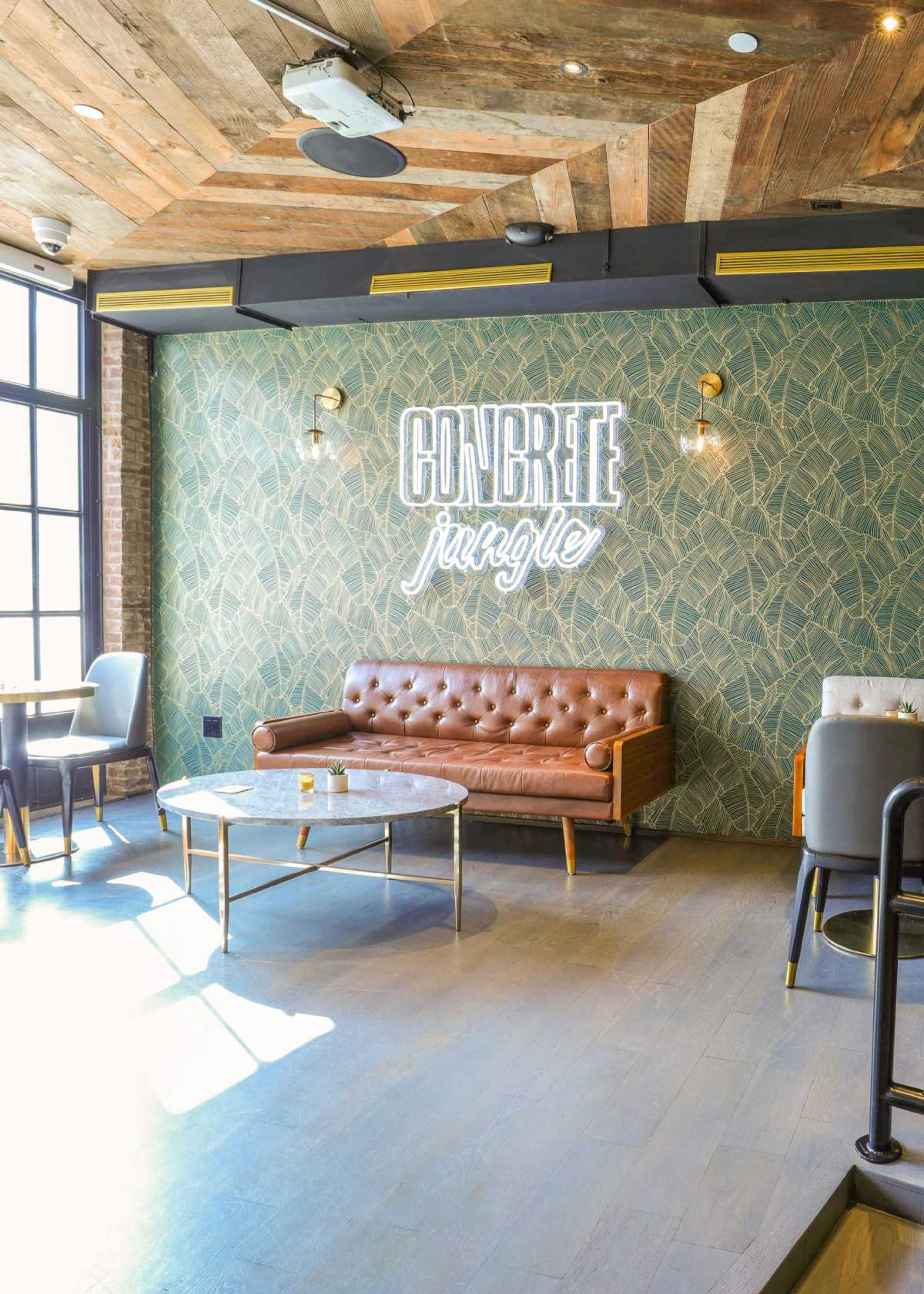 A stylish lounge area features a brown leather sofa, a round coffee table, and a neon sign that says "CONCRETE jungle" against a patterned green wall.