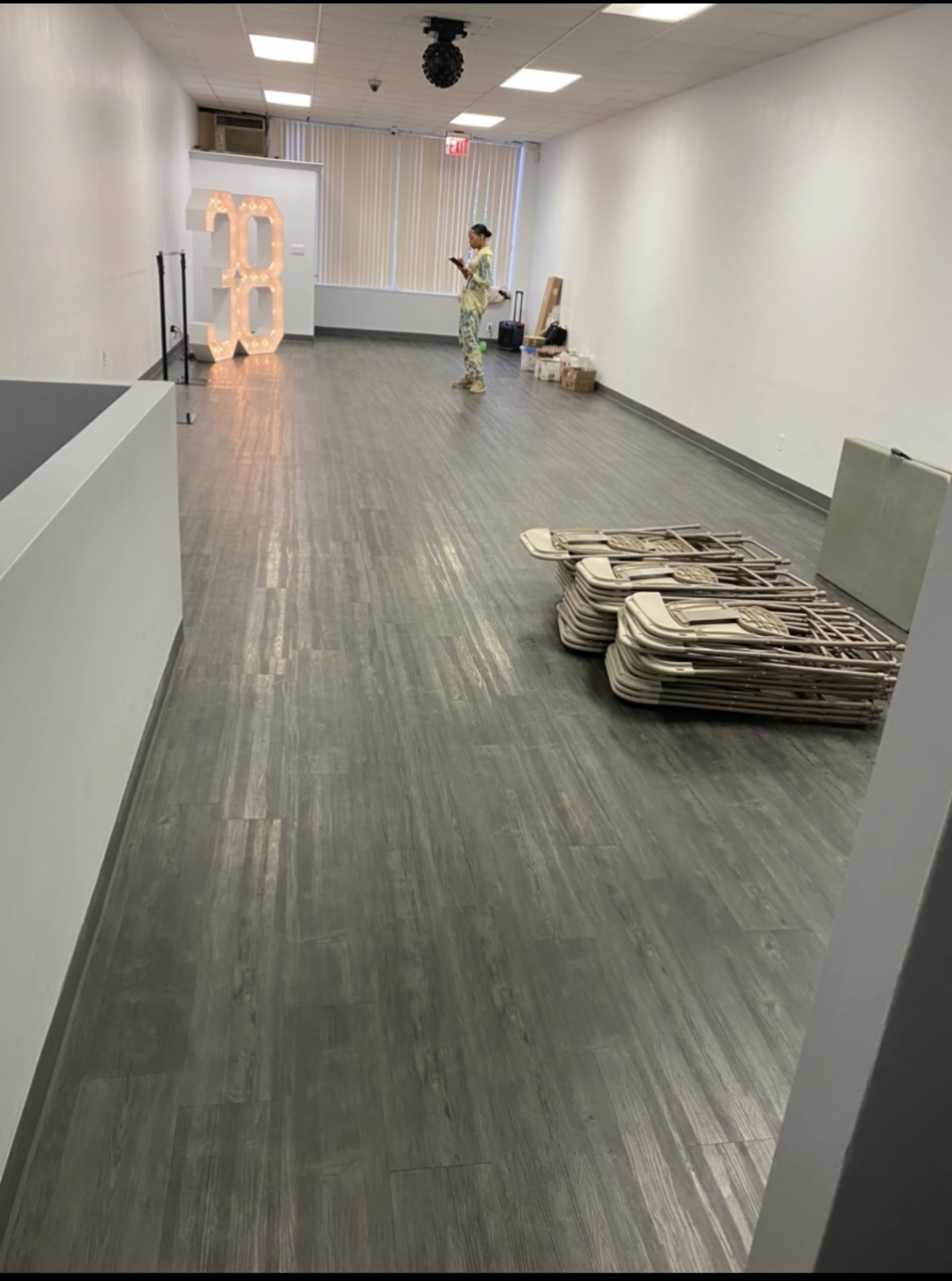 An empty room with gray flooring, a stack of folding chairs on the right, and a person standing in the background near illuminated numbers.
