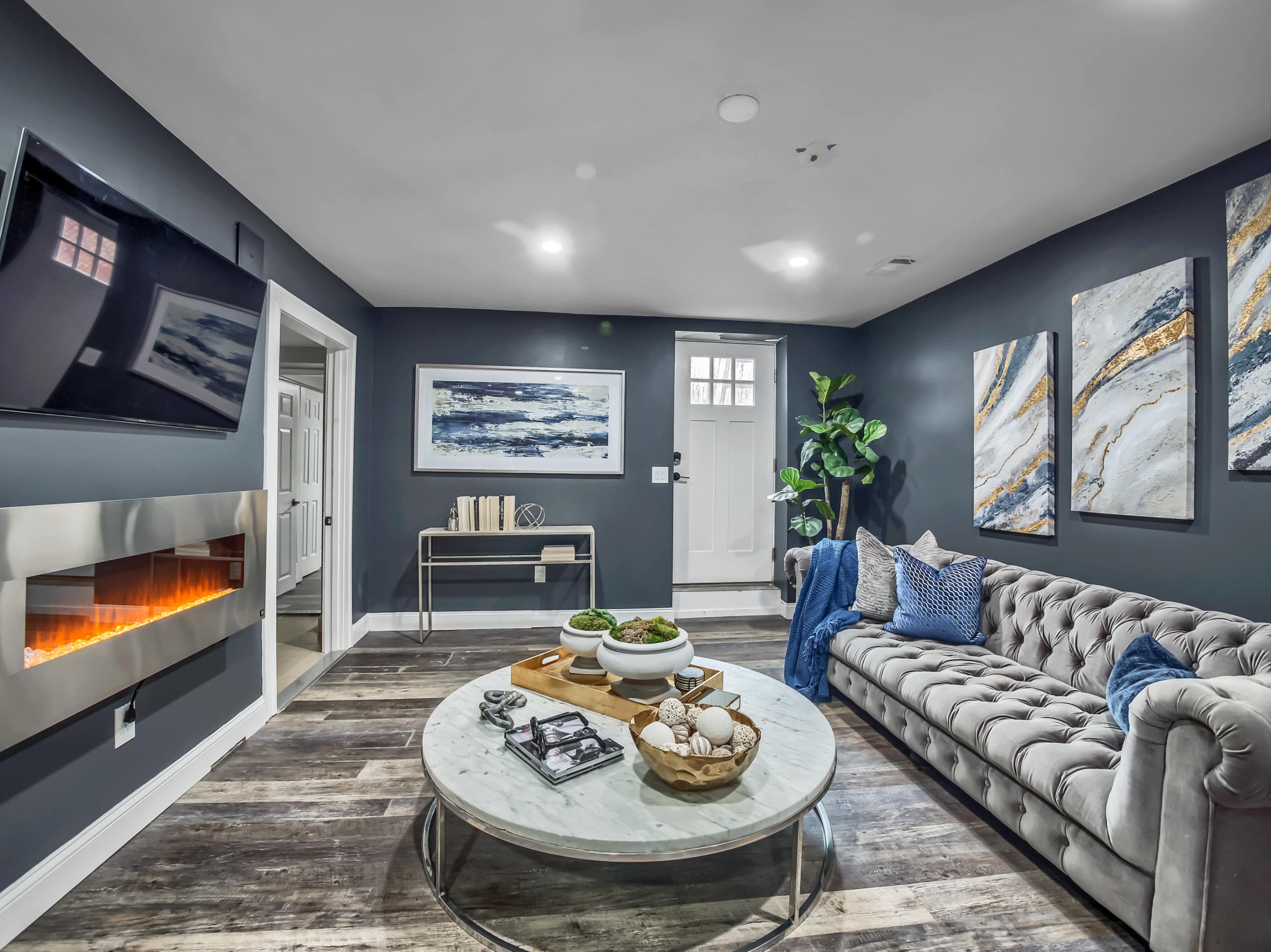 A modern living room with a gray tufted sofa, a round marble coffee table, a wall-mounted television, and an electric fireplace beneath a set of abstract paintings.