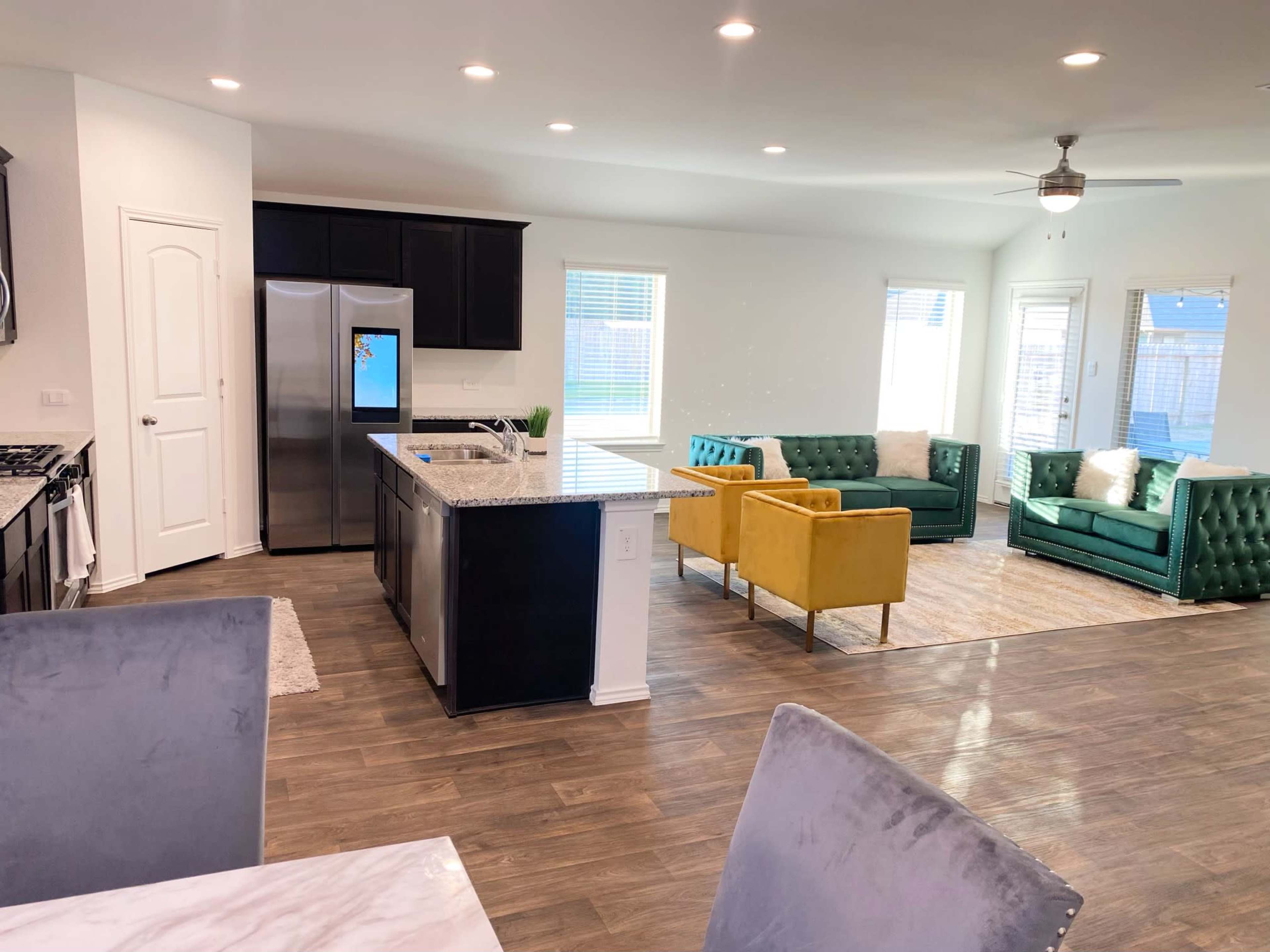 A modern open-concept living space features a kitchen with dark cabinetry and stainless steel appliances, adjacent to a bright sitting area with green and yellow sofas.