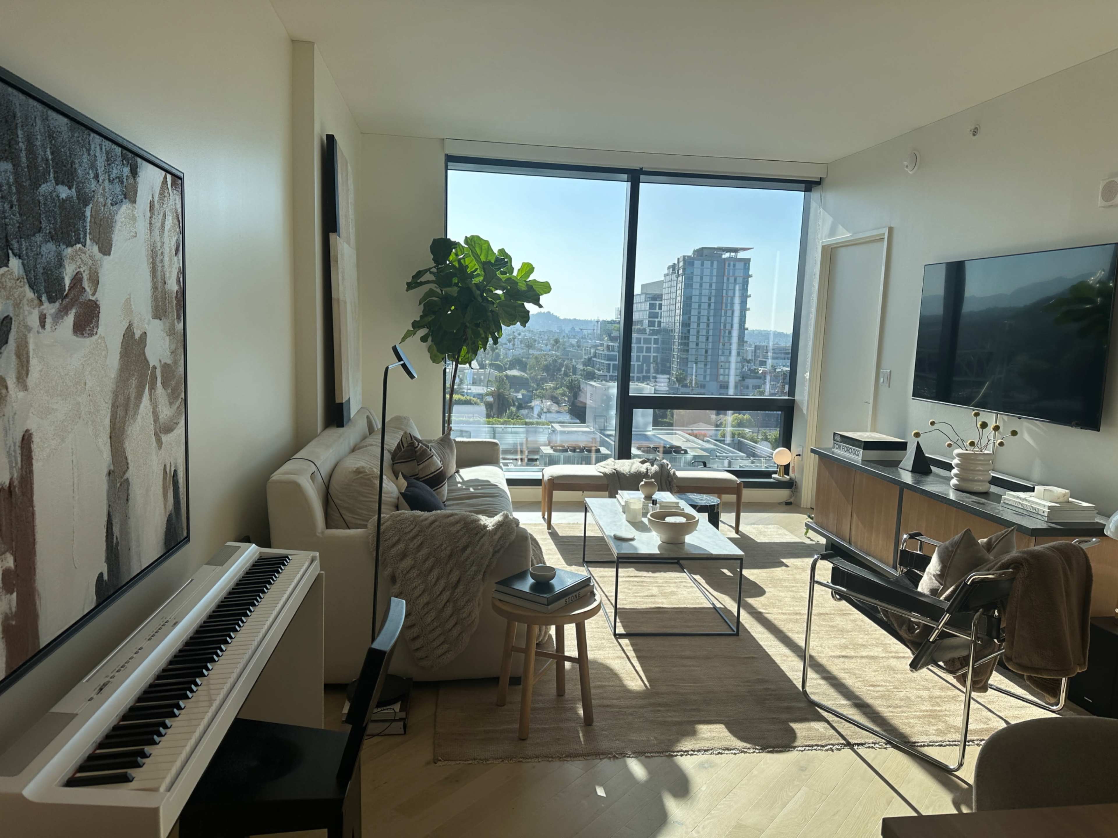 The image shows a modern living room with a large window overlooking a cityscape, a sofa, a coffee table, a keyboard, and various decorative elements.