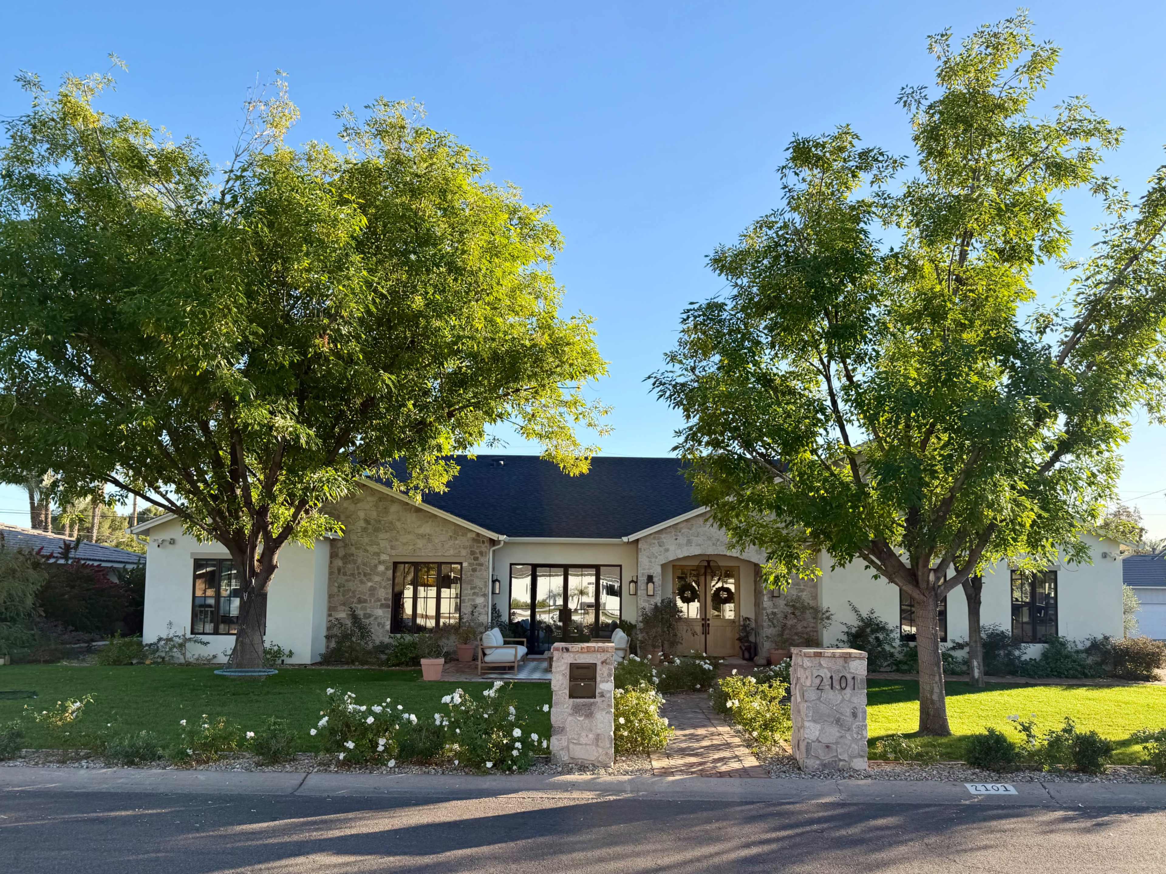 A single-story house with a stone facade and large windows is set on a grassy lawn with two large trees in front.