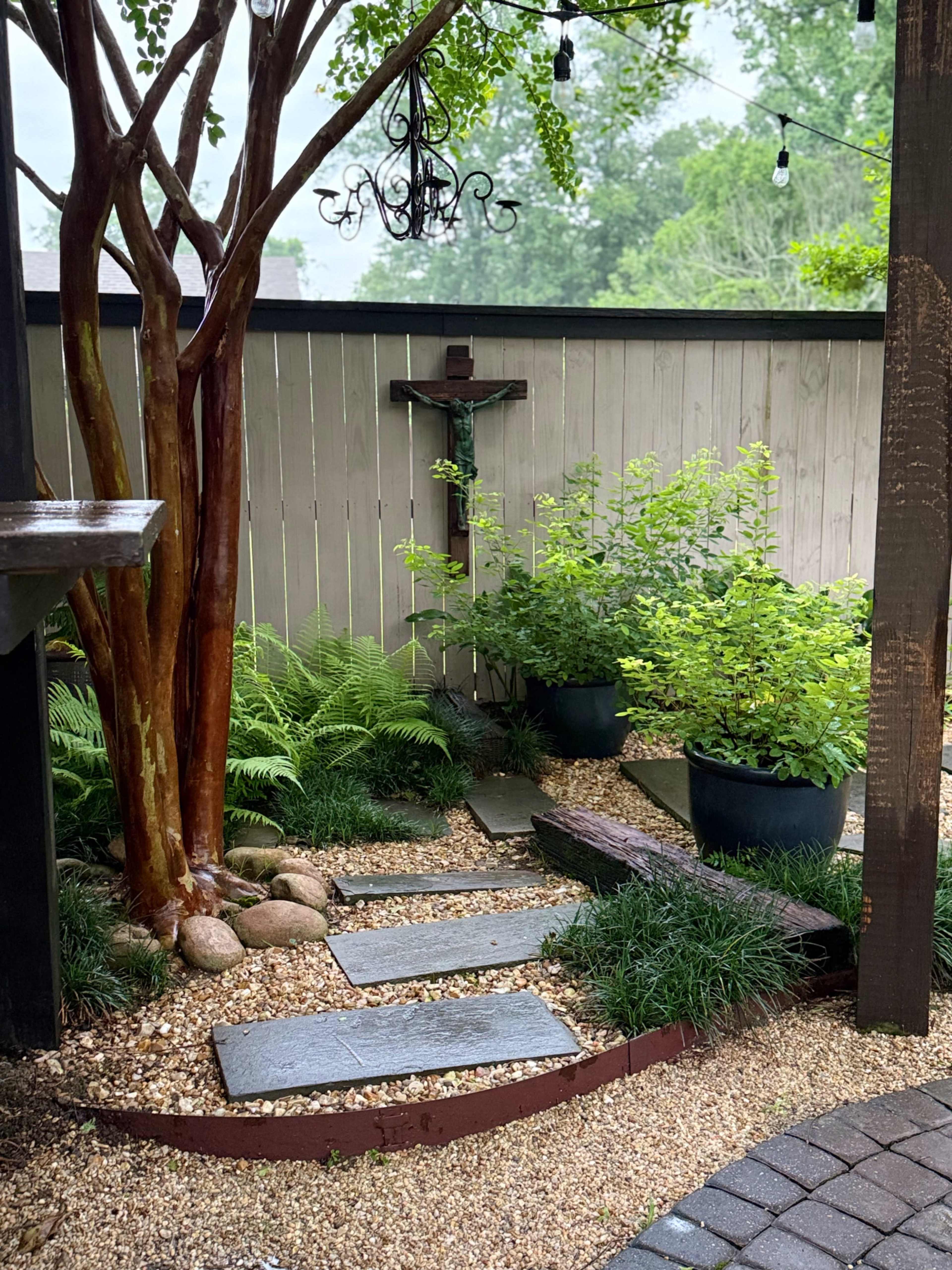 The image shows a landscaped garden area with stone pathways, a wooden cross on the wall, and greenery including ferns and potted plants.