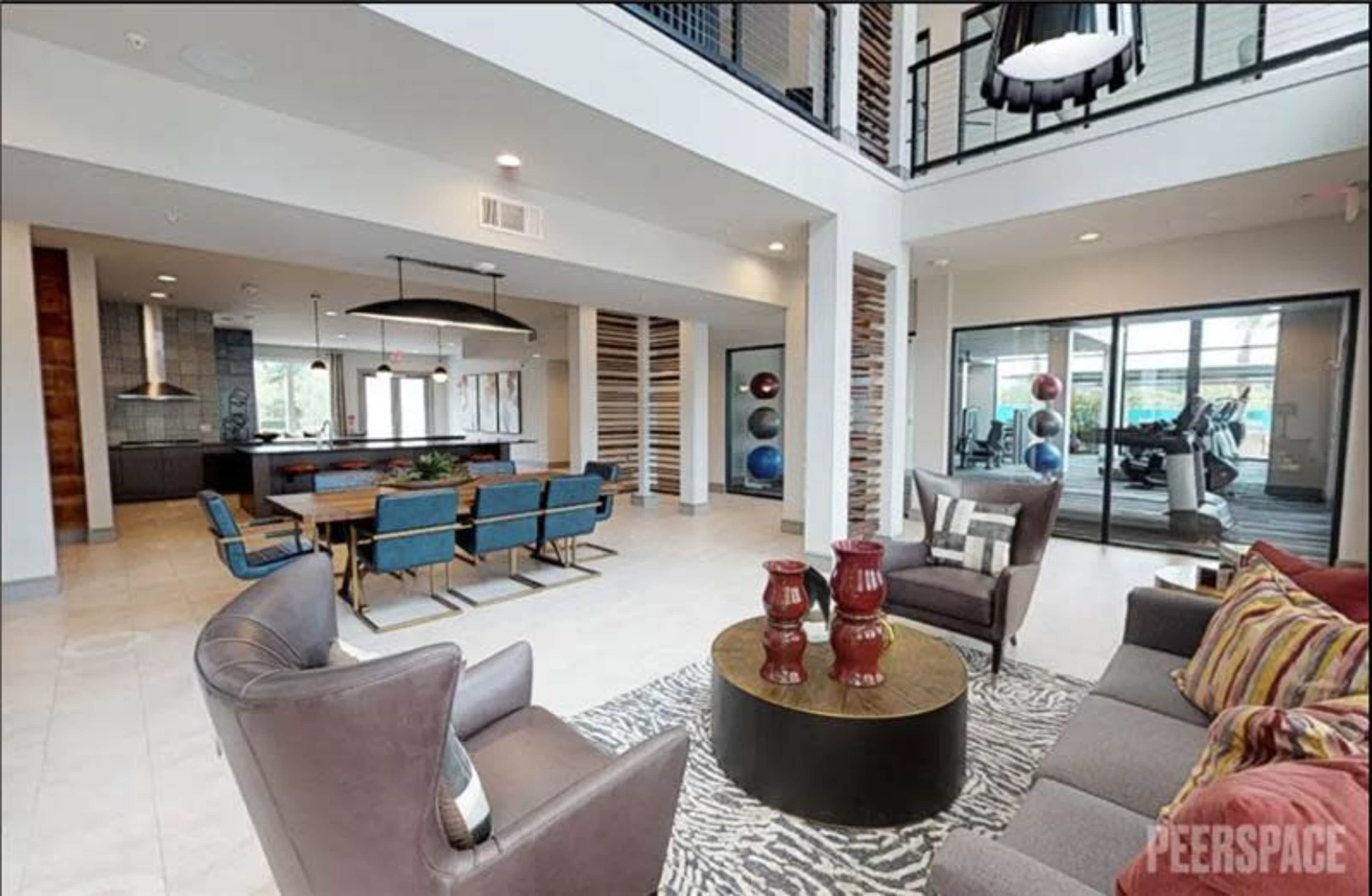 The image shows a modern open-concept living space with a seating area featuring two armchairs and a couch, a dining area with blue chairs, and a gym area visible through large glass doors.