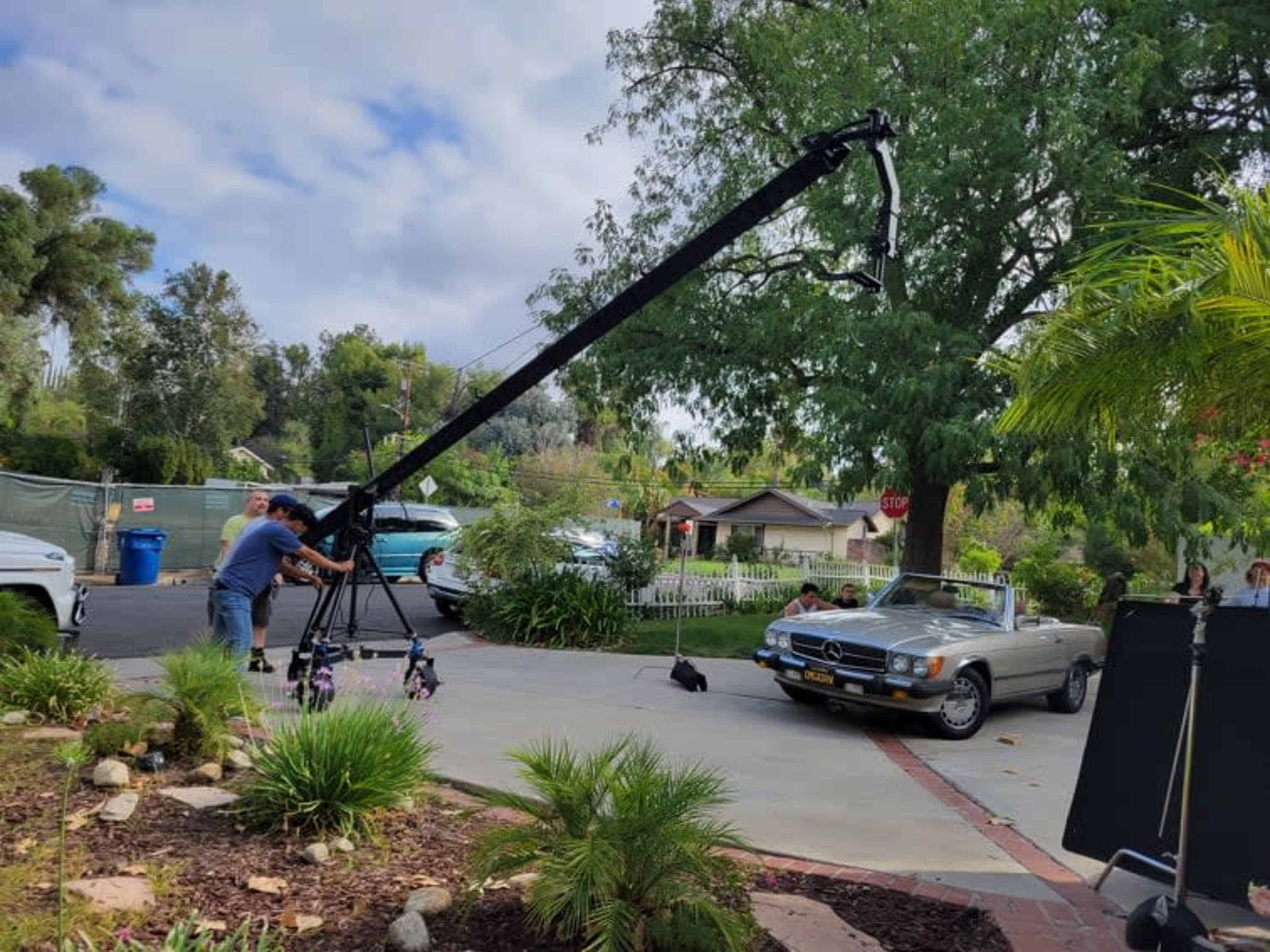 A camera crew operates a crane near a classic car in a residential area surrounded by greenery.