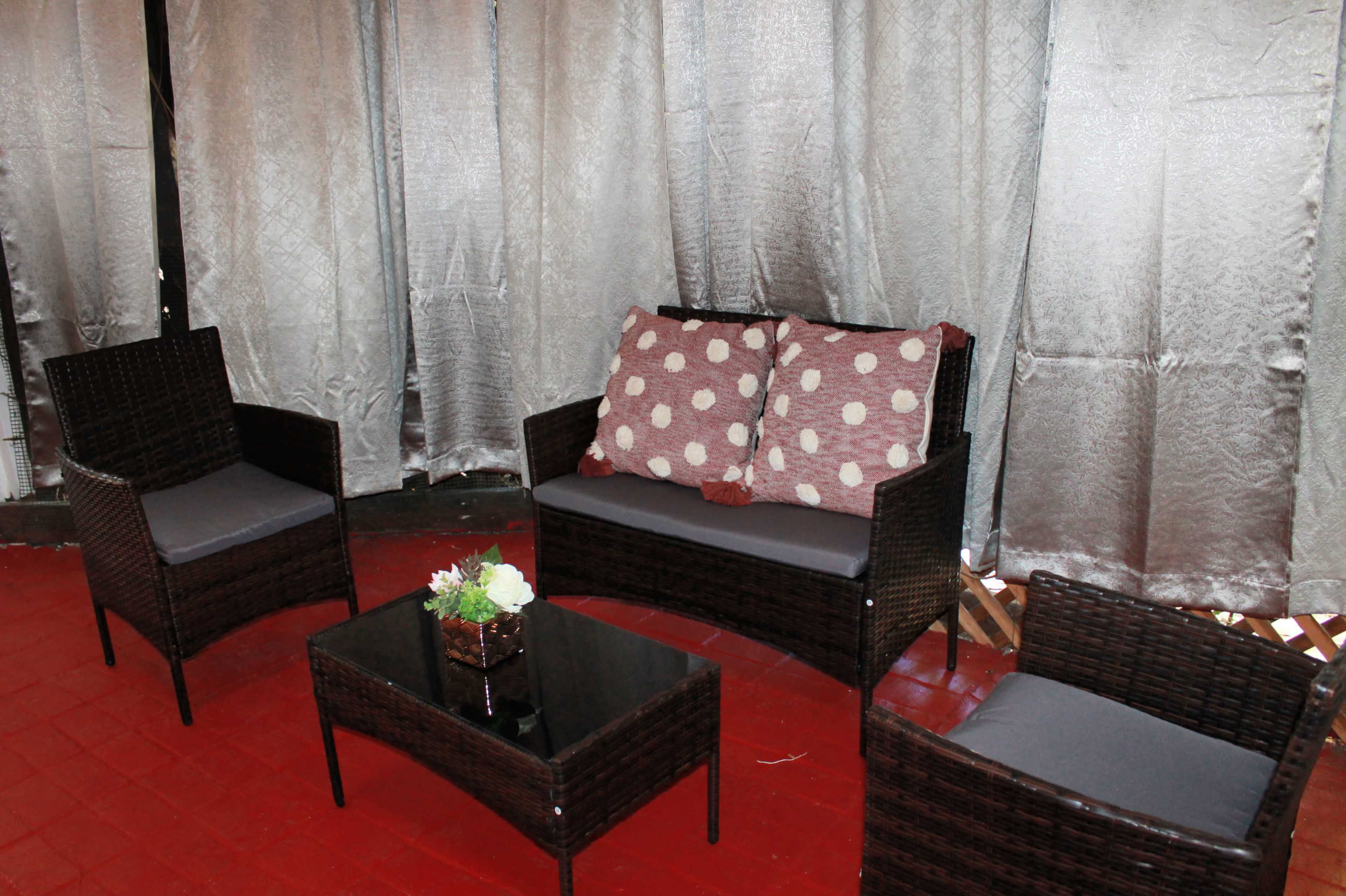 A seating area features a rectangular glass table surrounded by four woven wicker chairs and a two-seat sofa with polka dot cushions, set against a backdrop of grey curtains.