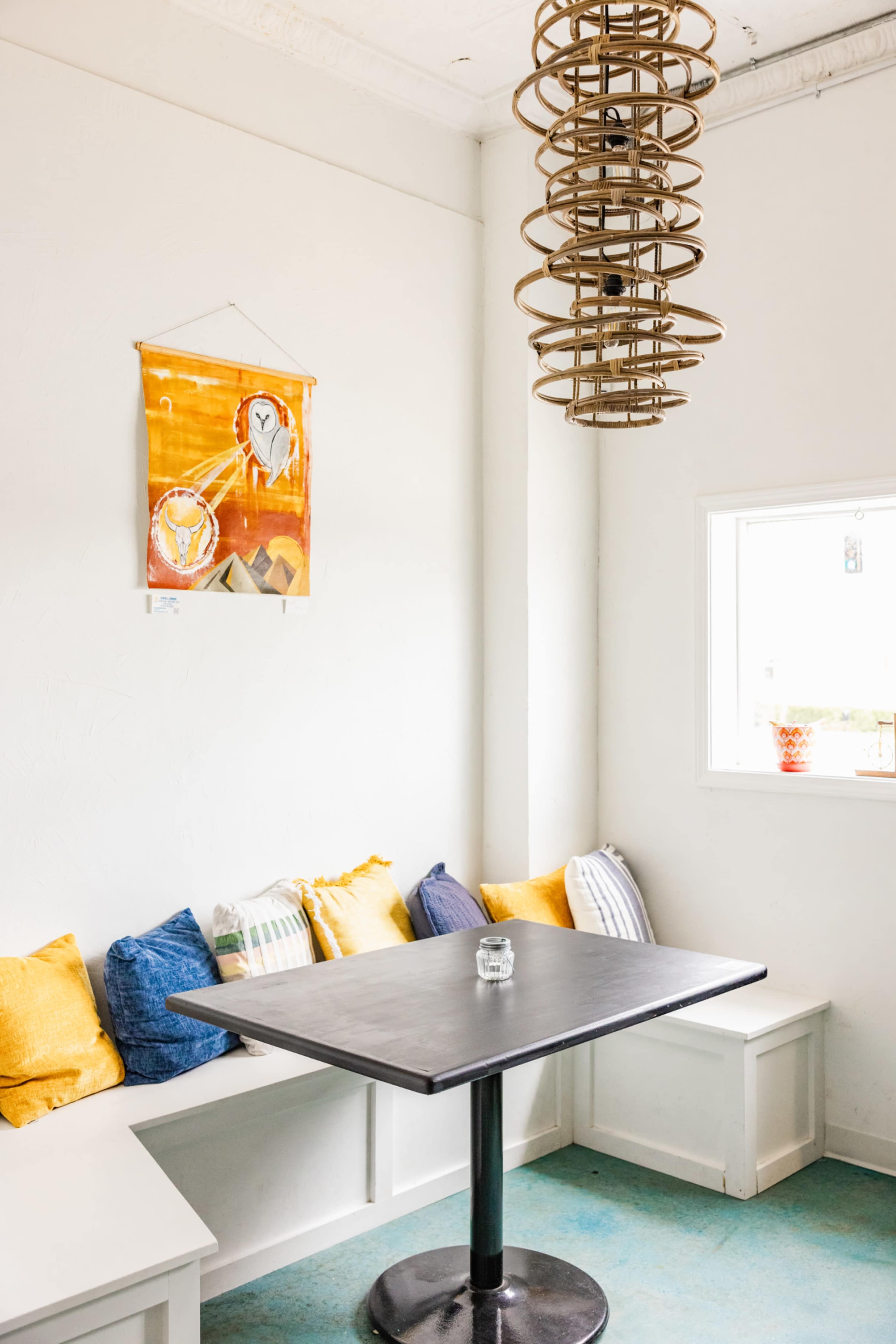 A cozy seating area features a table with a round base, surrounded by colorful cushions, under a decorative hanging light and a wall-mounted artwork.