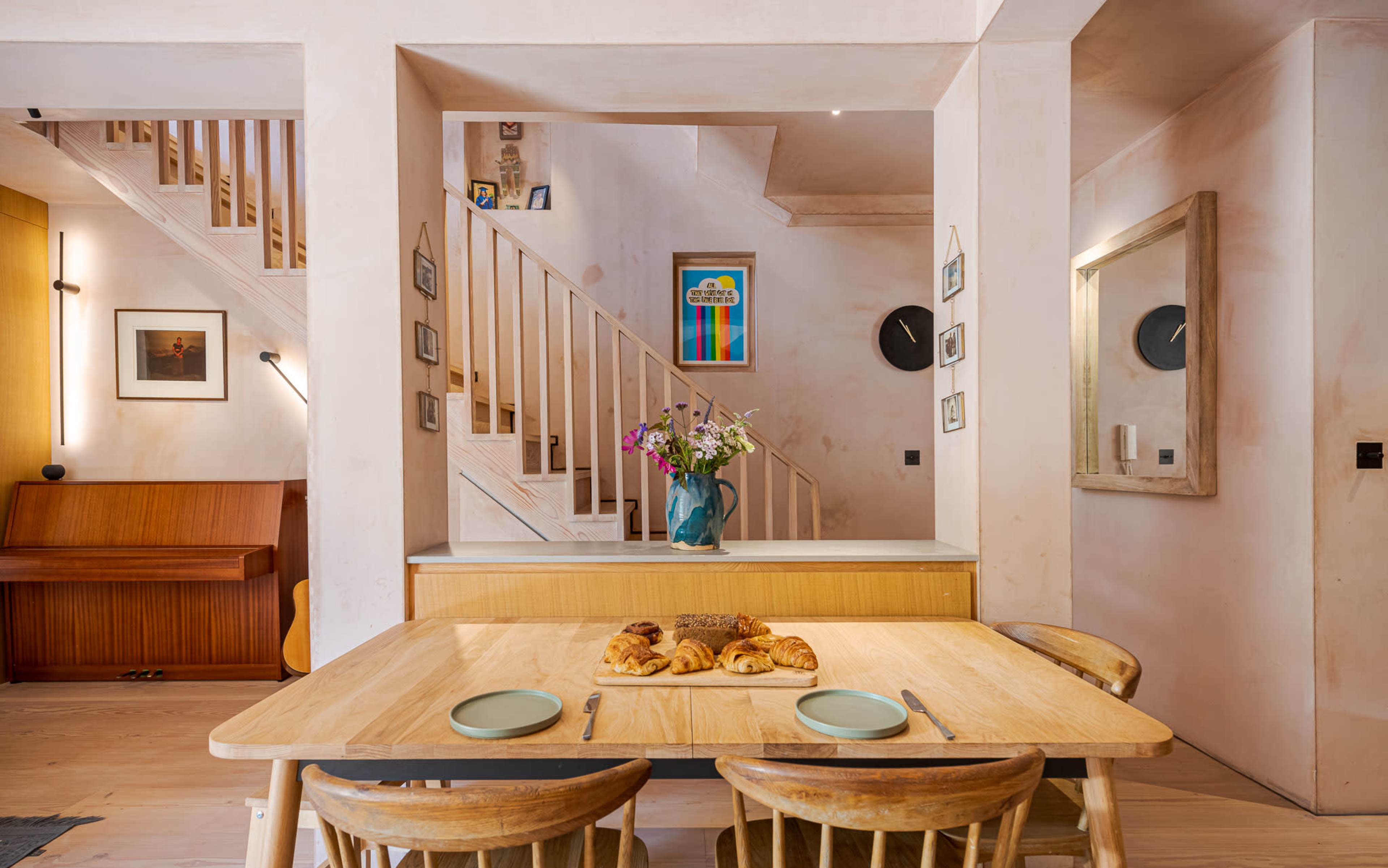 The image shows a dining area with a wooden table set for breakfast, featuring croissants and plates, facing a staircase with a colorful framed artwork on the wall.