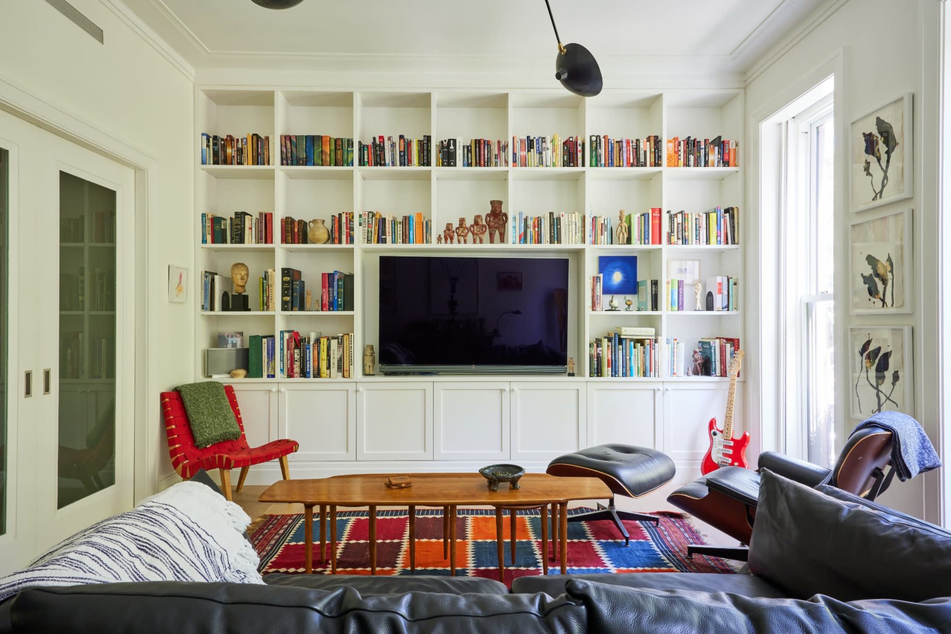 A modern living room features a large bookshelf filled with books, a television mounted on a white wall, and comfortable seating arranged around a patterned area rug.