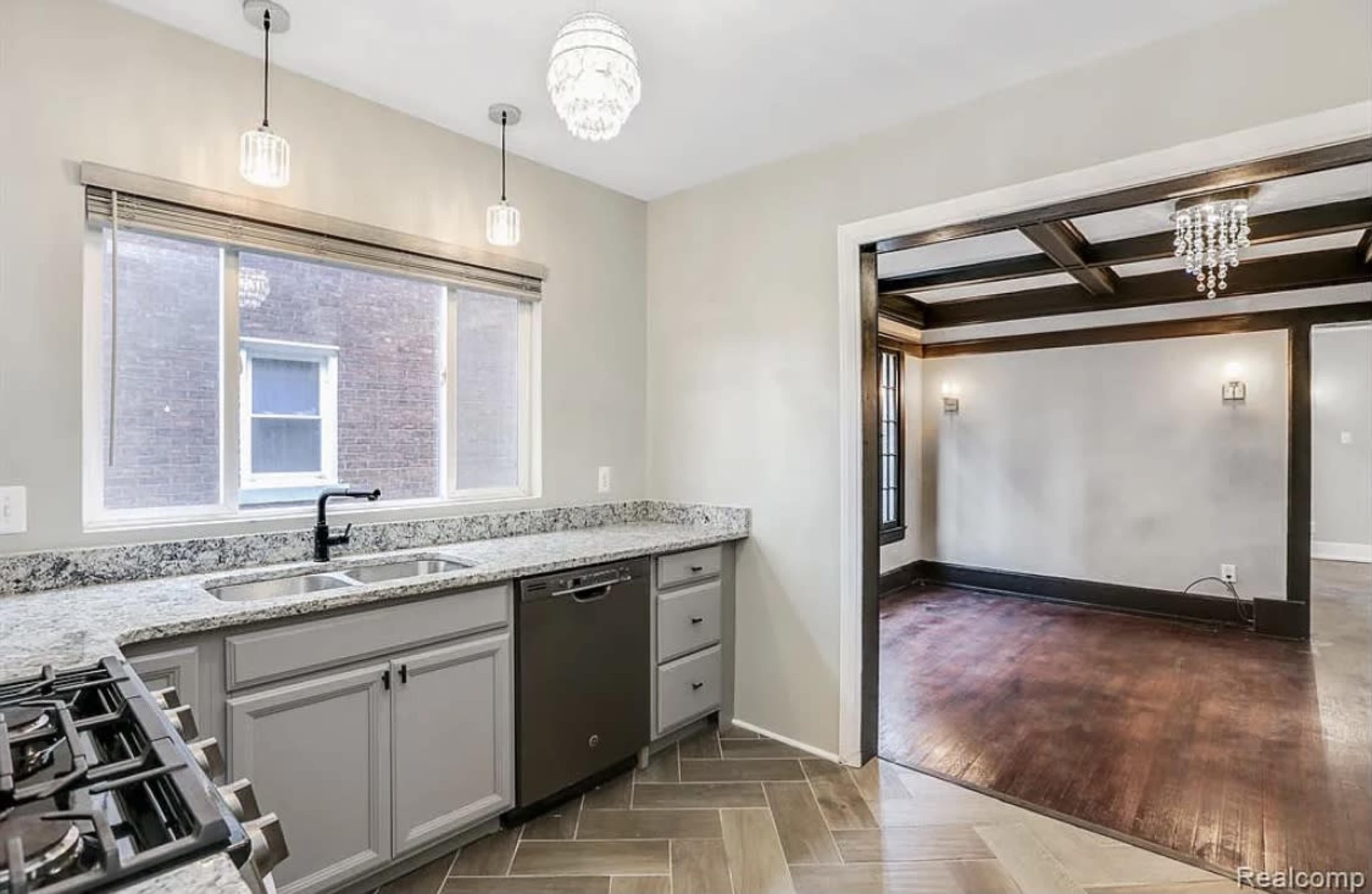 A modern kitchen area with granite countertops, a double sink, a dishwasher, and a view into a living room featuring wooden beams and a chandelier.