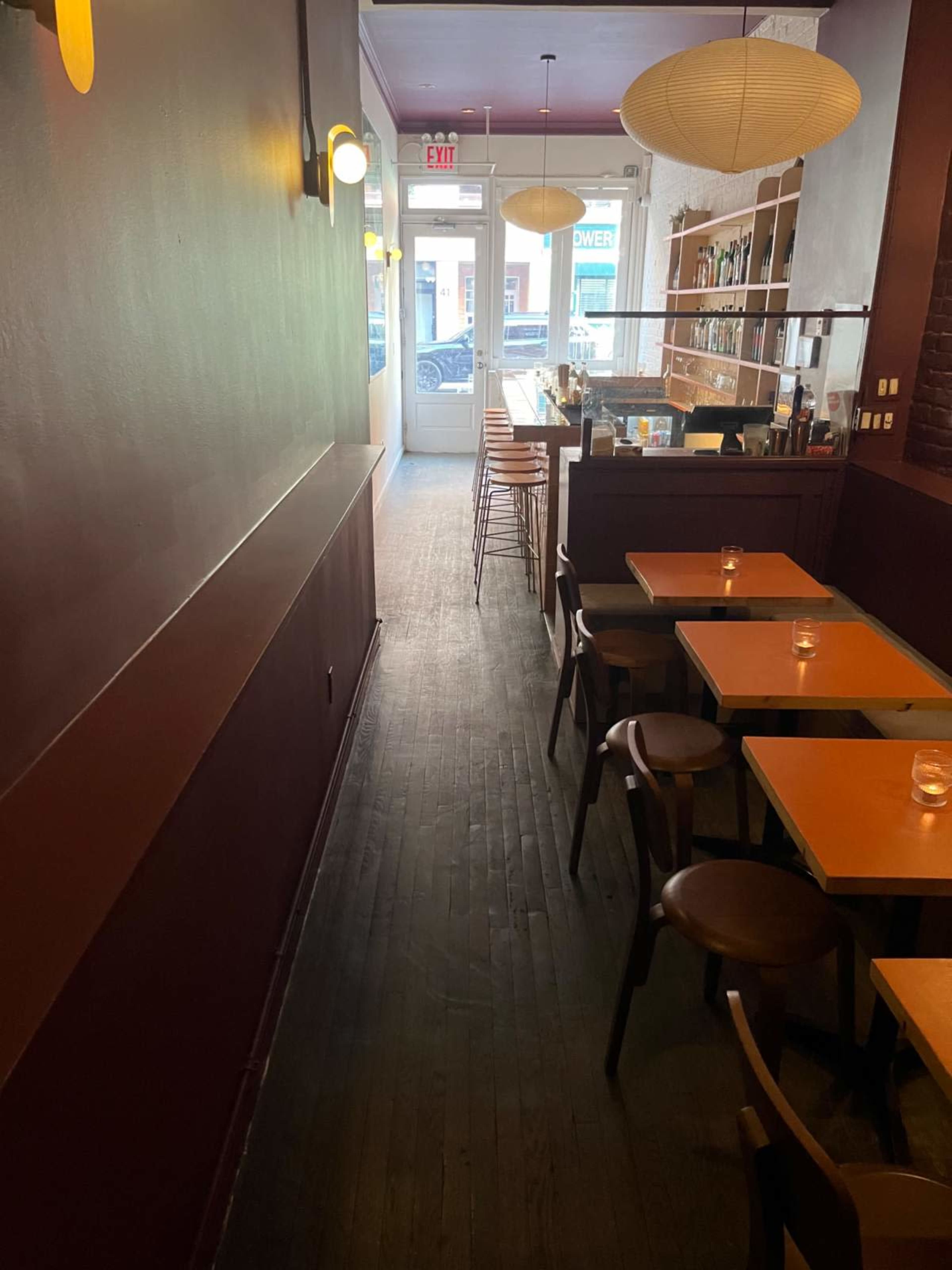 The image shows a narrow restaurant interior with wooden tables and chairs, illuminated by soft lighting, leading to a glass entrance door.