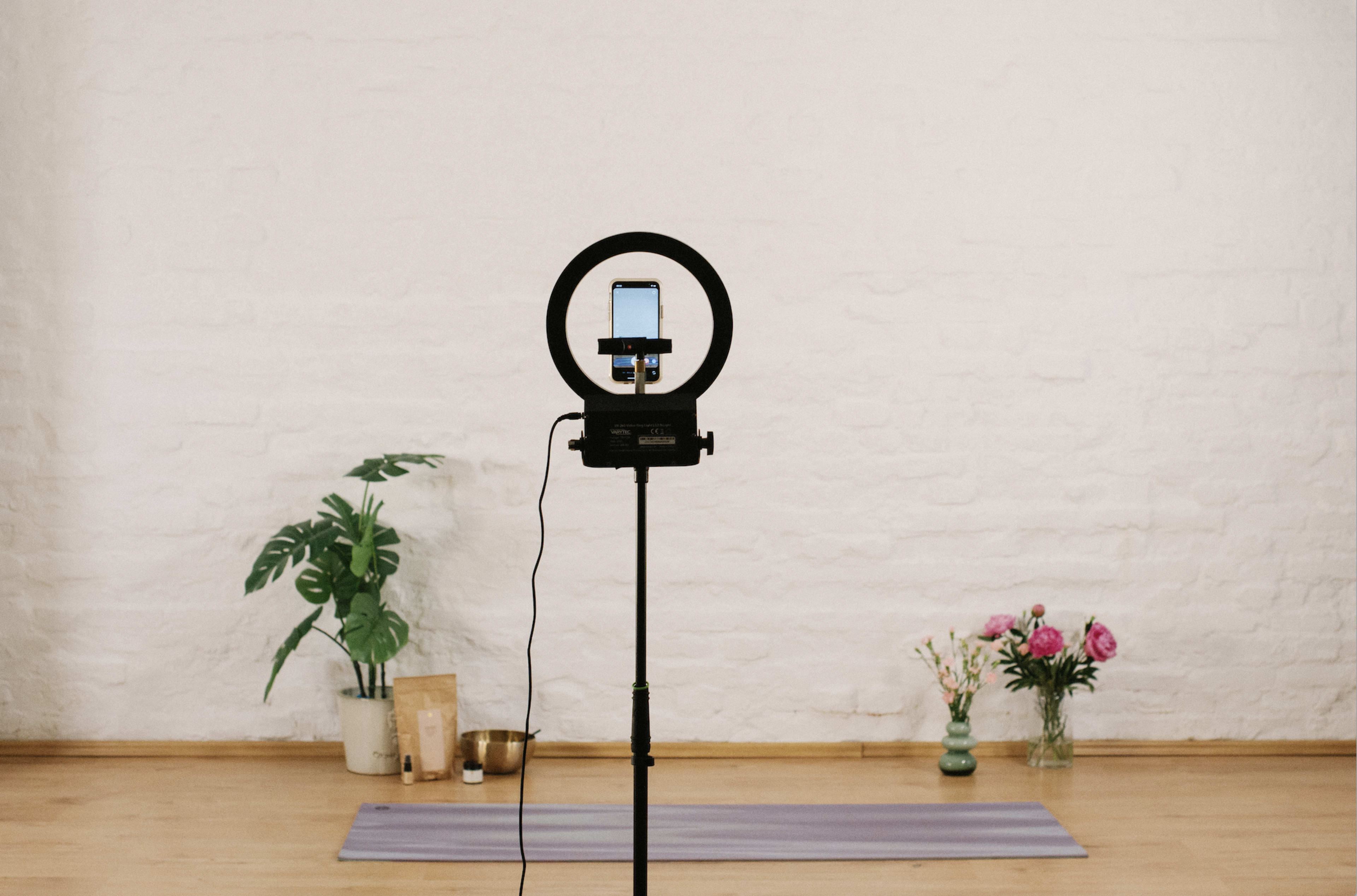 A ring light mounted on a stand holds a phone facing a yoga mat in a minimalist room with plants and flowers in the background.